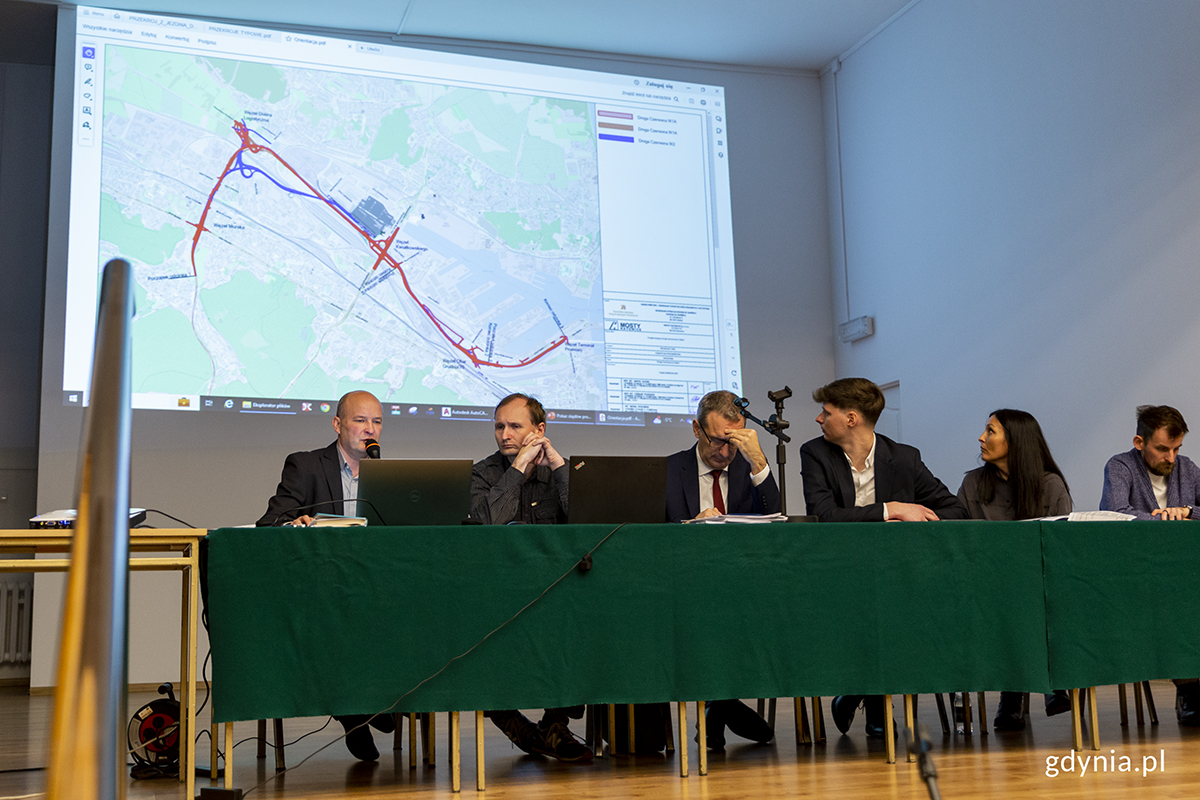 Kilka os&oacute;b siedzi za stołem, za nimi ekran z prezentacja.