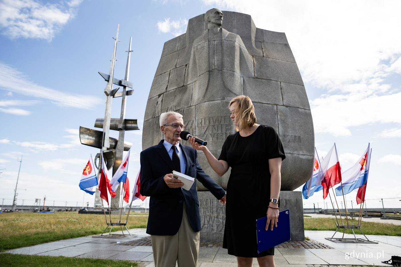Kobieta i mężczyzna stoją pod pomnikiem. Mężczyzna przemawia, kobieta trzyma mikrofon.