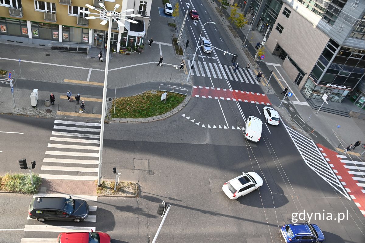 Skrzyżowanie w widoku z g&oacute;ry, widoczne kolorowe samochody i piesi oraz ściany okolicznych budynk&oacute;w 