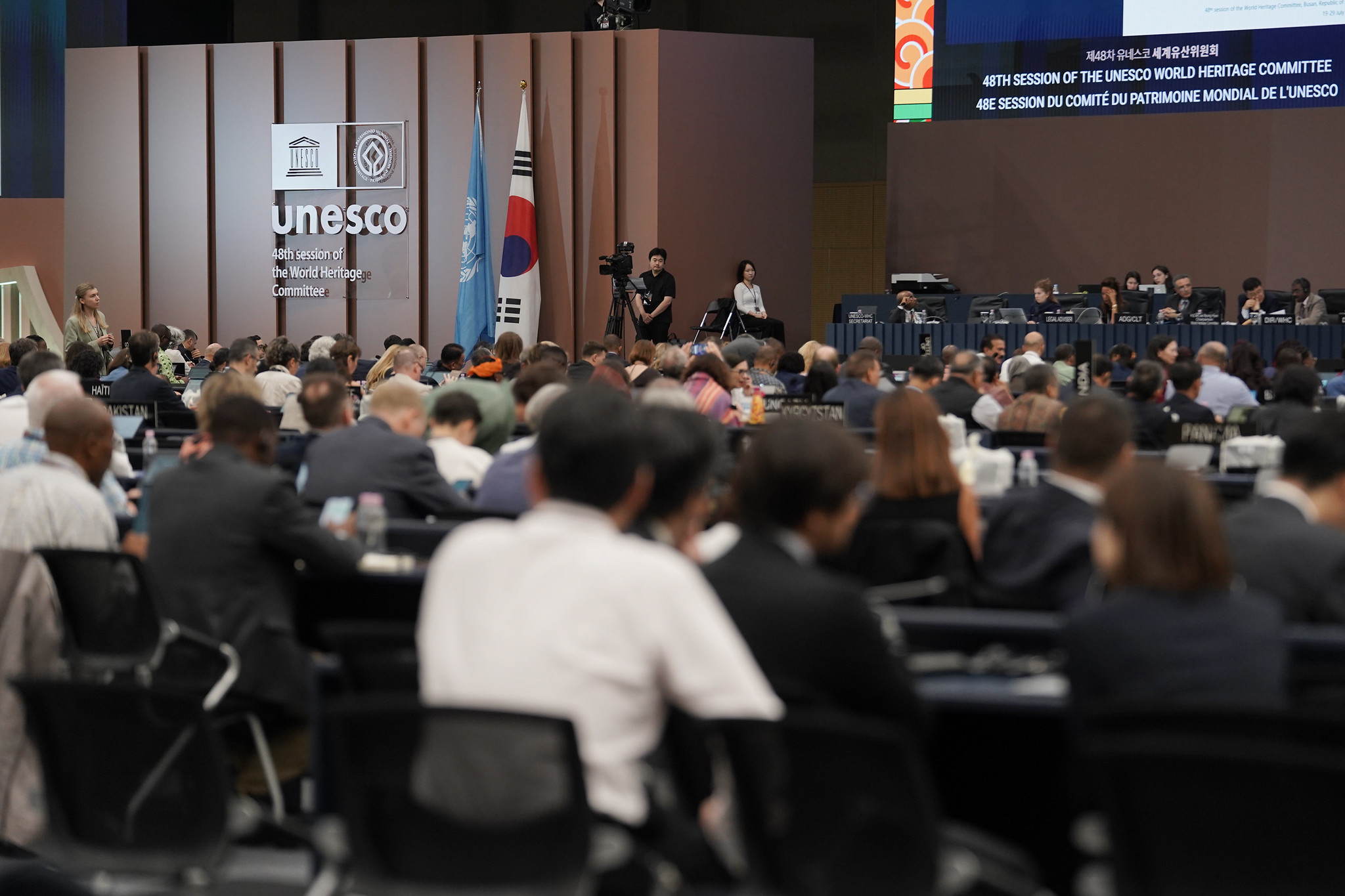 Sala konferencyjna, tłum ludzi przy stolikach, logotyp 48. sesji Komitetu Światowego Dziedzictwa UNESCO