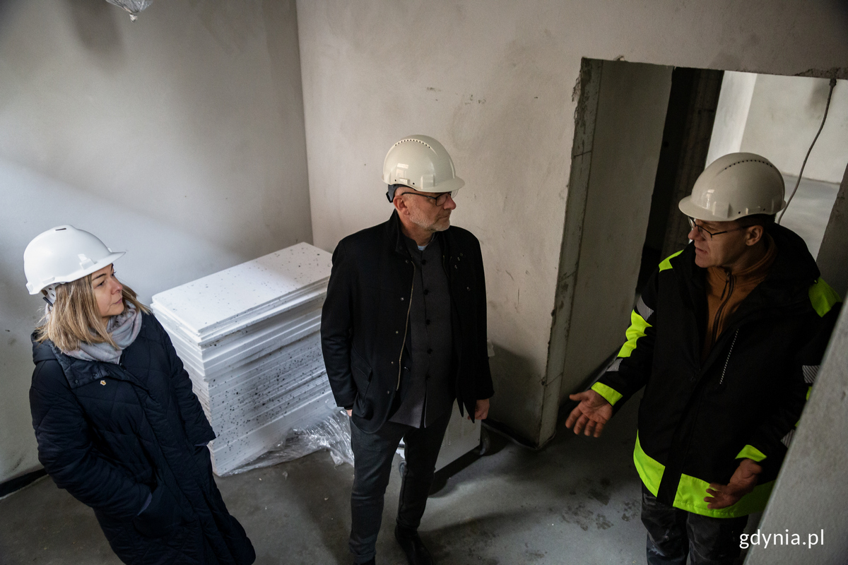 Dw&oacute;ch mężczyzn i kobieta w kaskach rozmawiają wewnątrz budynku na budowie.