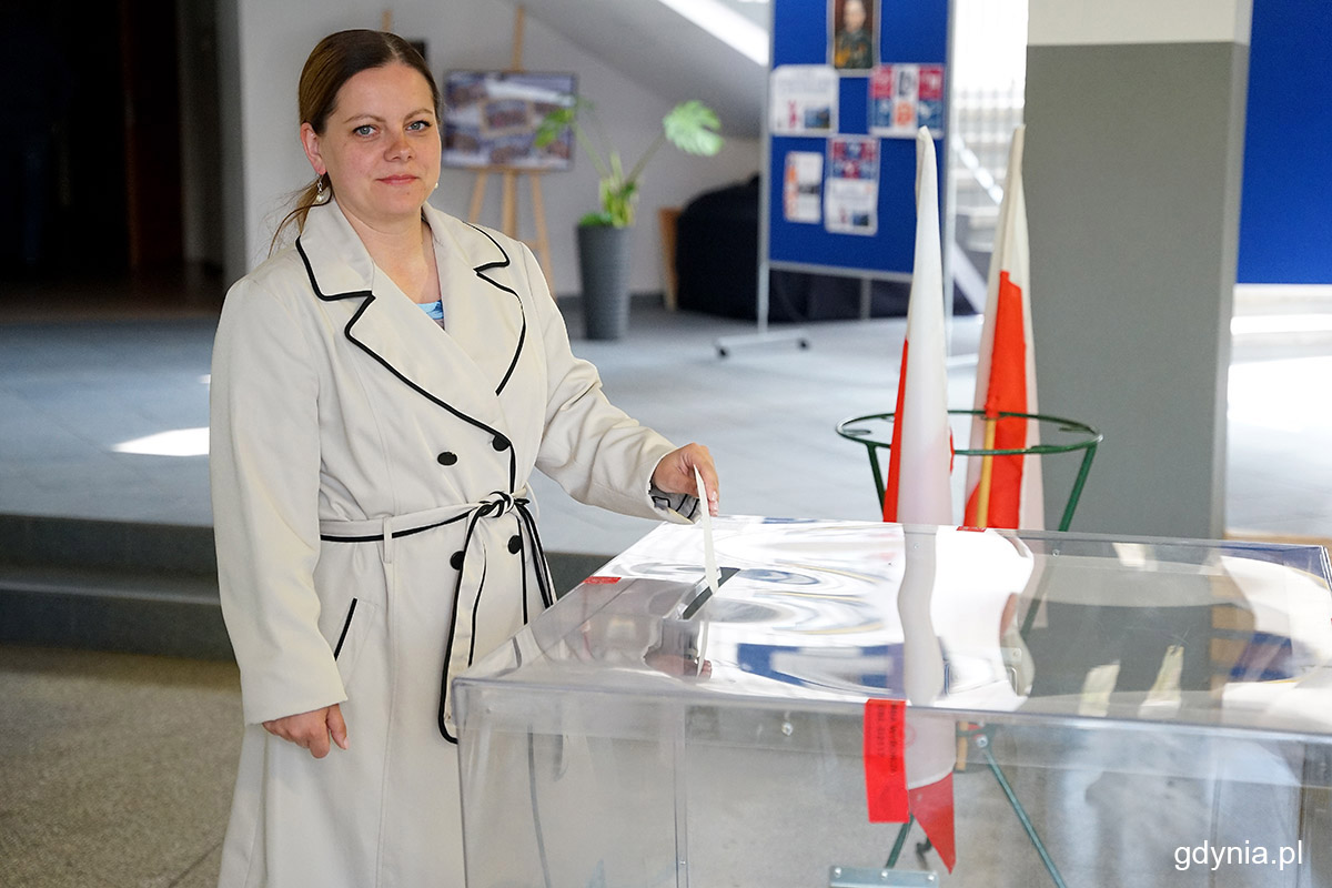 Aleksandra Kosiorek pozuje do zdjęcia przy urnie wyborczej, urna, głos w rękach, w tle hol szkolny