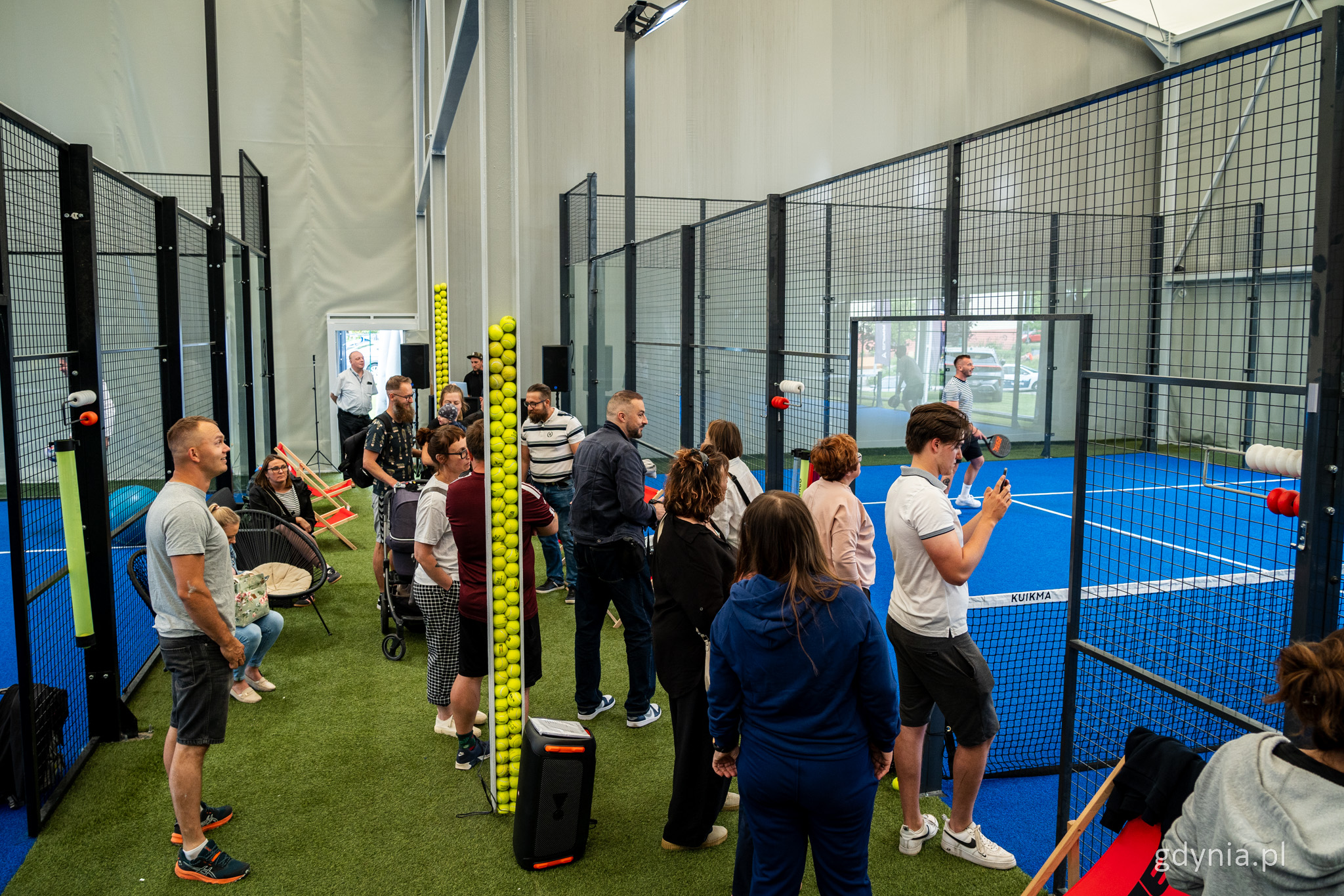 Kort do gry w padla, niebieska nawierzchnia, przejście między kortami, kilkanaście os&oacute;b stoi między kortami