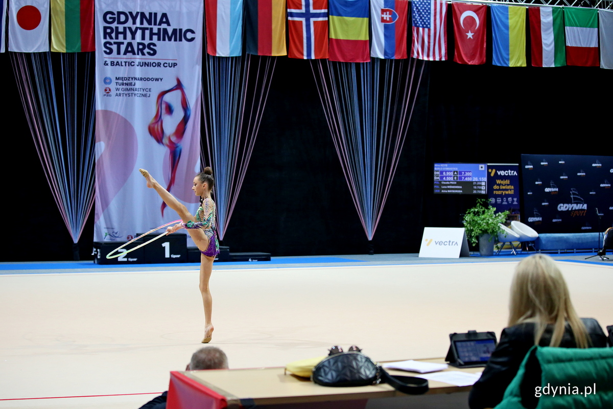 Na zdjęciu zawodniczka podczas układu, w tle flagi państw, kt&oacute;re występują w zawodach, rozmyta na pierwszym planie ekipa sędziowska.