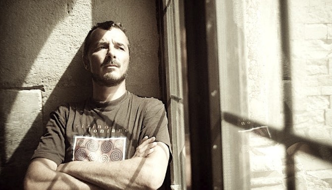 Grzegorz Franczak - nominowany do Nagrody Literackiej GDYNIA w kategorii przekład na język polski - w jasnym t-shircie z nadrukiem. Opiera się o ścianę i ma założone ręce // fot. Chiara Paiano