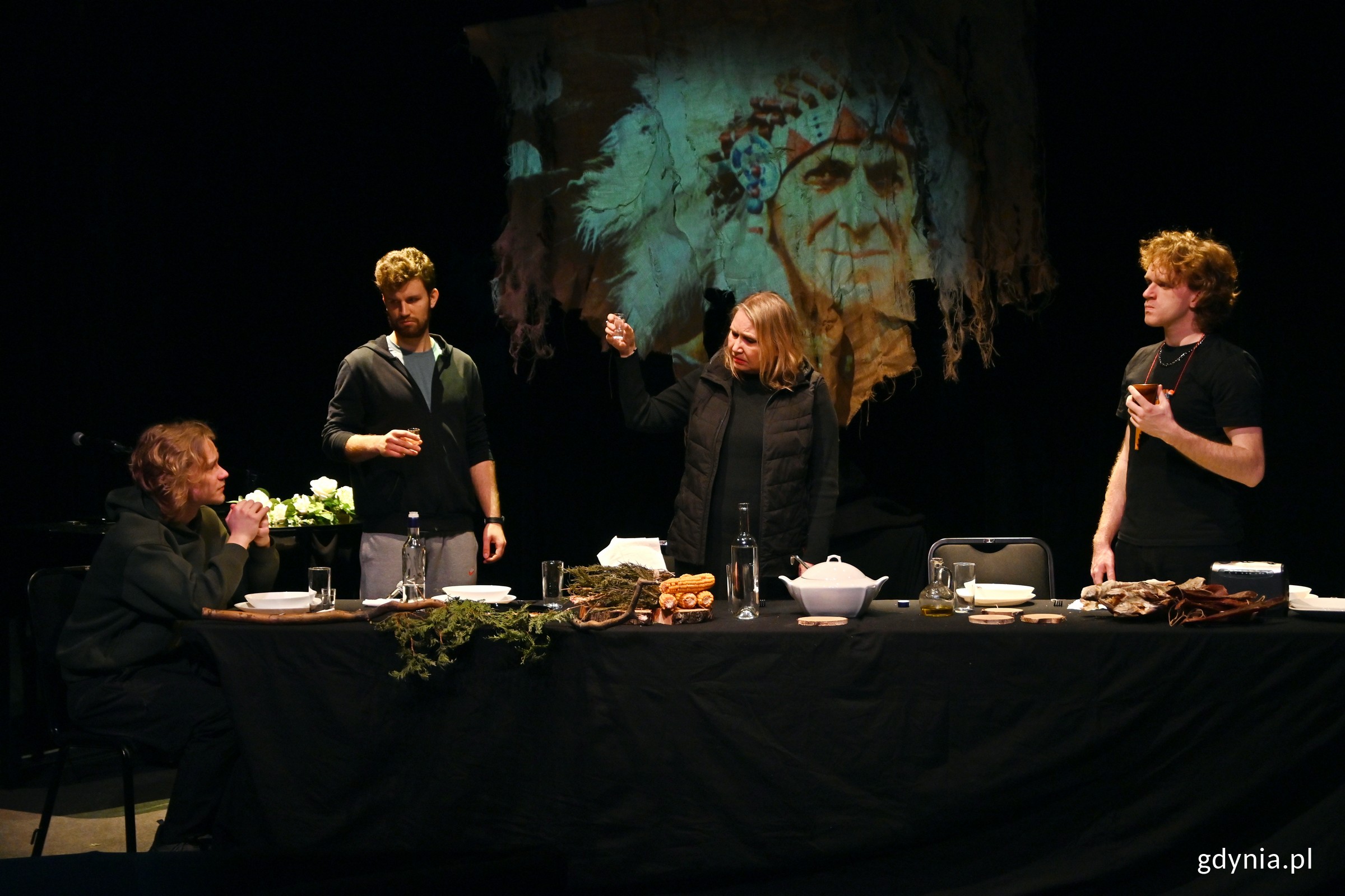 Zdjęcie z pr&oacute;by spektaklu &bdquo;Sat-Okh&rdquo; w reżyserii Aleksandry Skorupy. Scena zbiorowa. Aktorzy stojący i siedzący za stołem (fot. Magdalena Czernek)