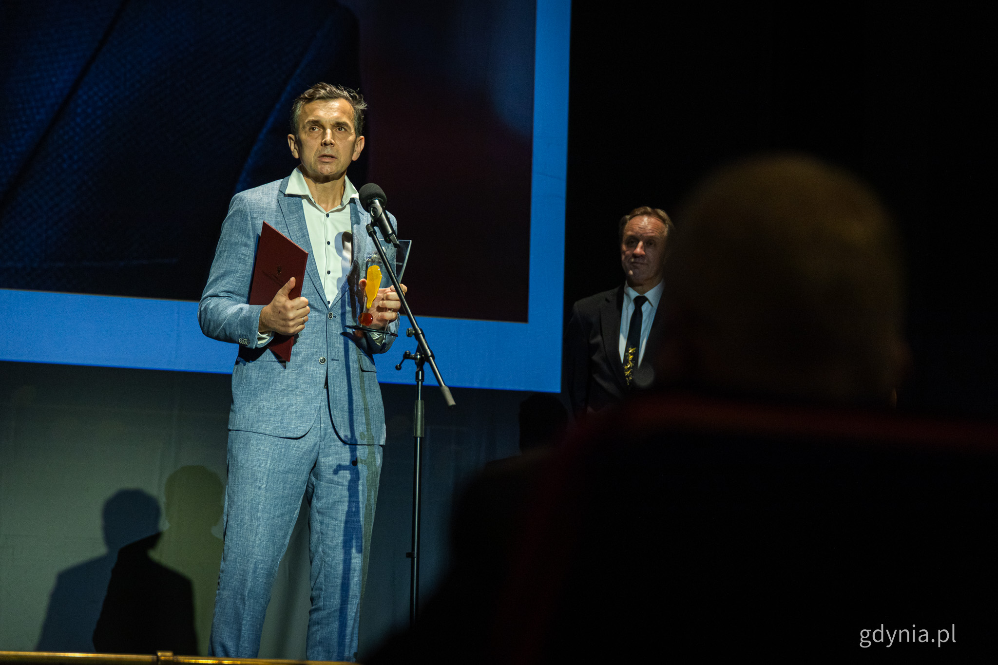Jacek Debis na scenie, trzyma statuetkę, przemawia do mikrofonu