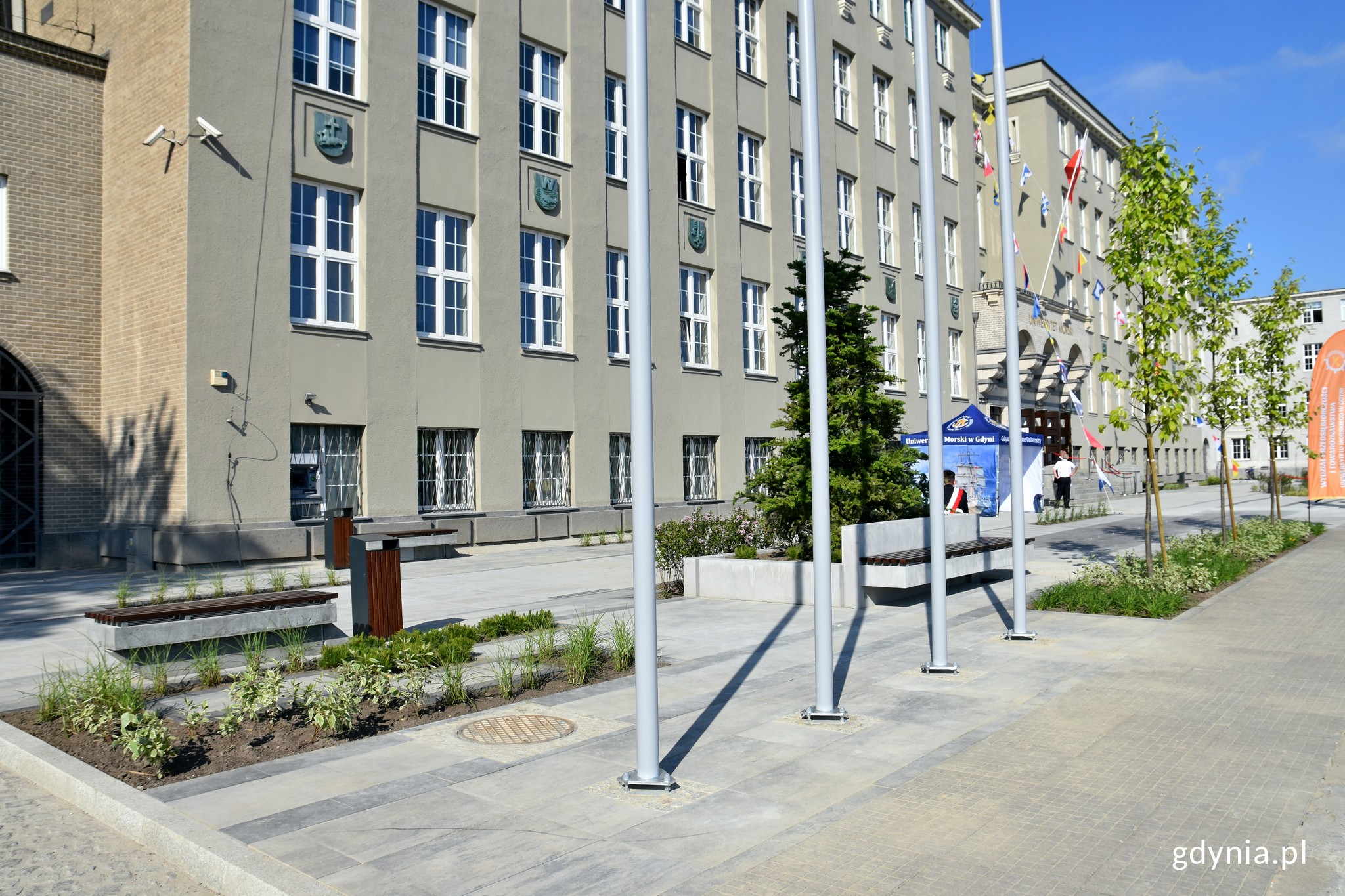 Przestrzeń reprezentacyjno-parkowa przed budynkiem Uniwersytetu Morskiego w Gdyni przy ul. Morskiej. Na pierwszym planie ławka i fragment zieleni - drzew i krzew&oacute;w posadzonych przed uczelnią. W tle mury budynku // fot. Magdalena Czernek