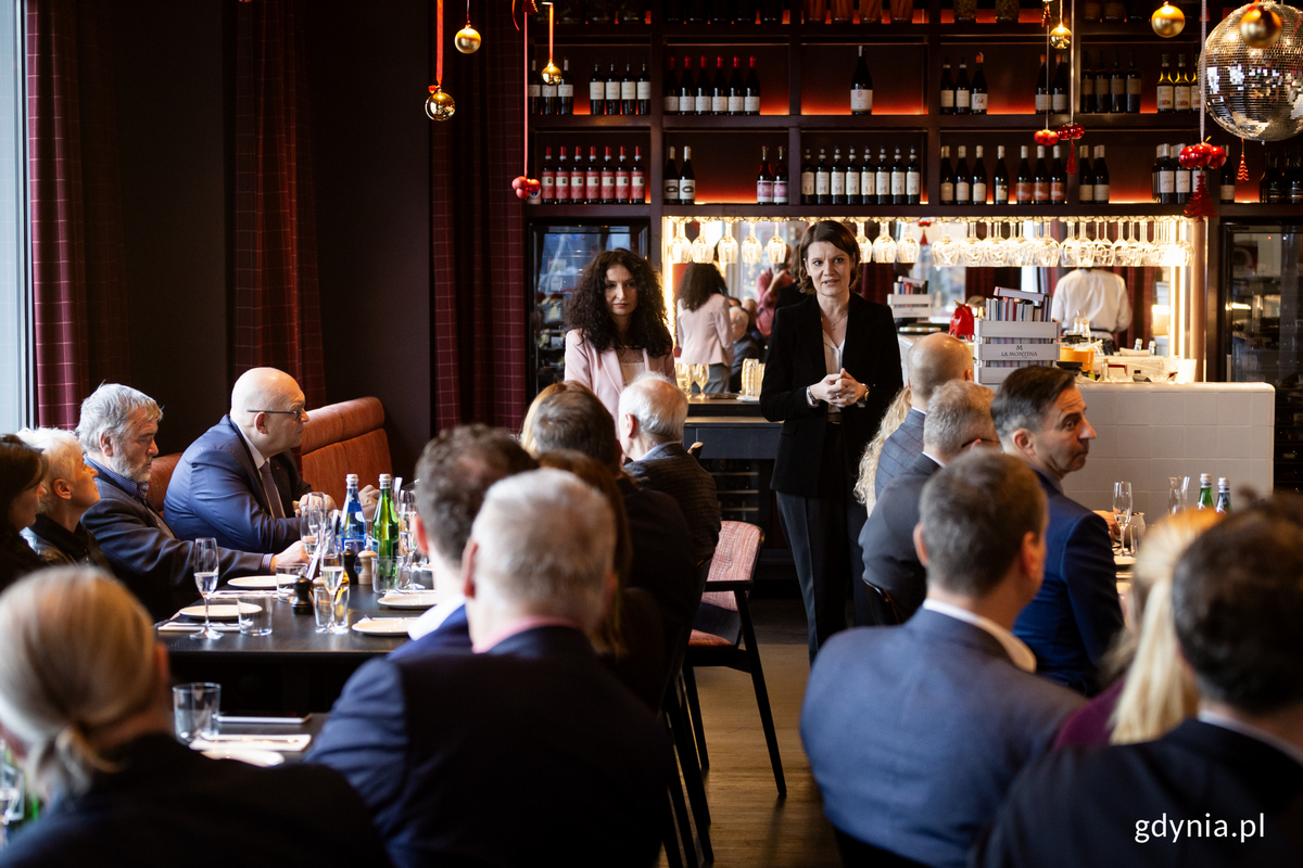 Dwie kobiety stoja i m&oacute;wia przed tłumem ludzi siedzących w restauracji.