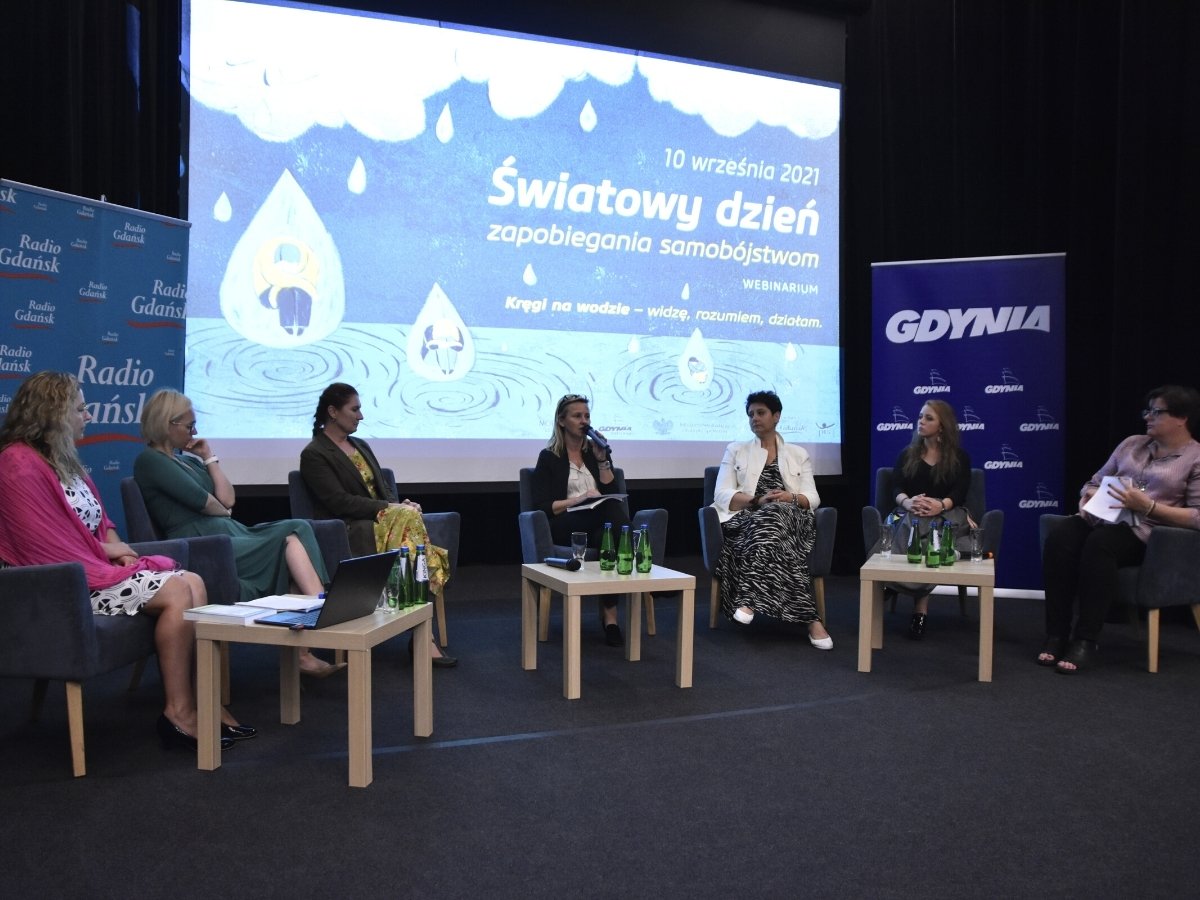 Grupa kobiet siedząca przy stolikach podczas konferencji. Jedna z nich trzyma mikrofon i przemawia. W tle bannery promocyjne kampanii.