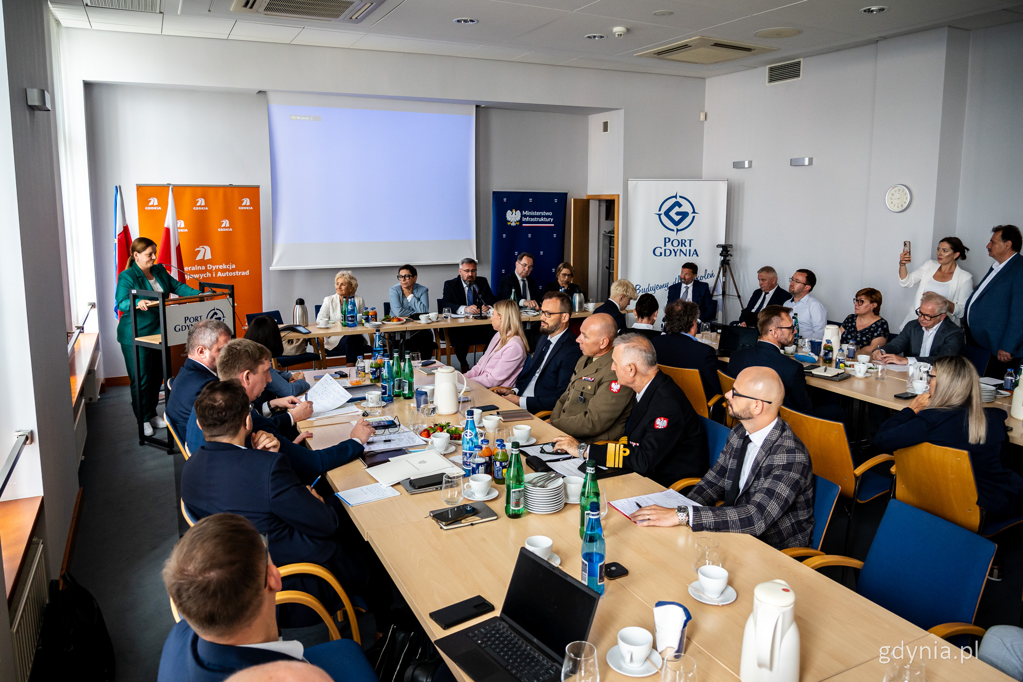 Sala konferencyjna, grupa os&oacute;b siedząca przy stołach, na środku stoly prezydium, ekran, przy m&oacute;wnicy w rogu Aleksandra Kosiorek