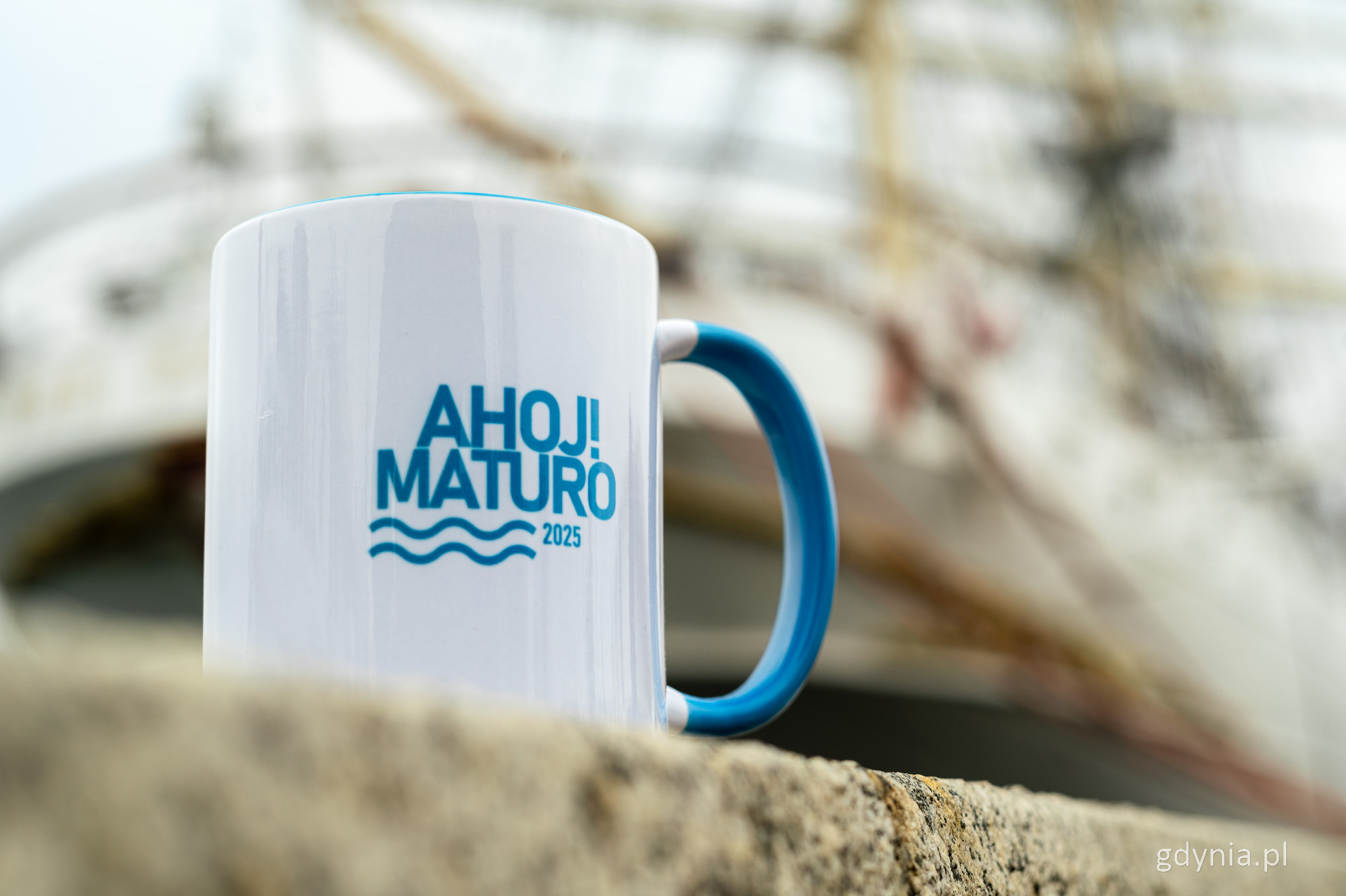 Biały kubek z niebieskim napisem "Ahoj! Matura" na tle Daru Pomorza, stoi na nabrzeżu