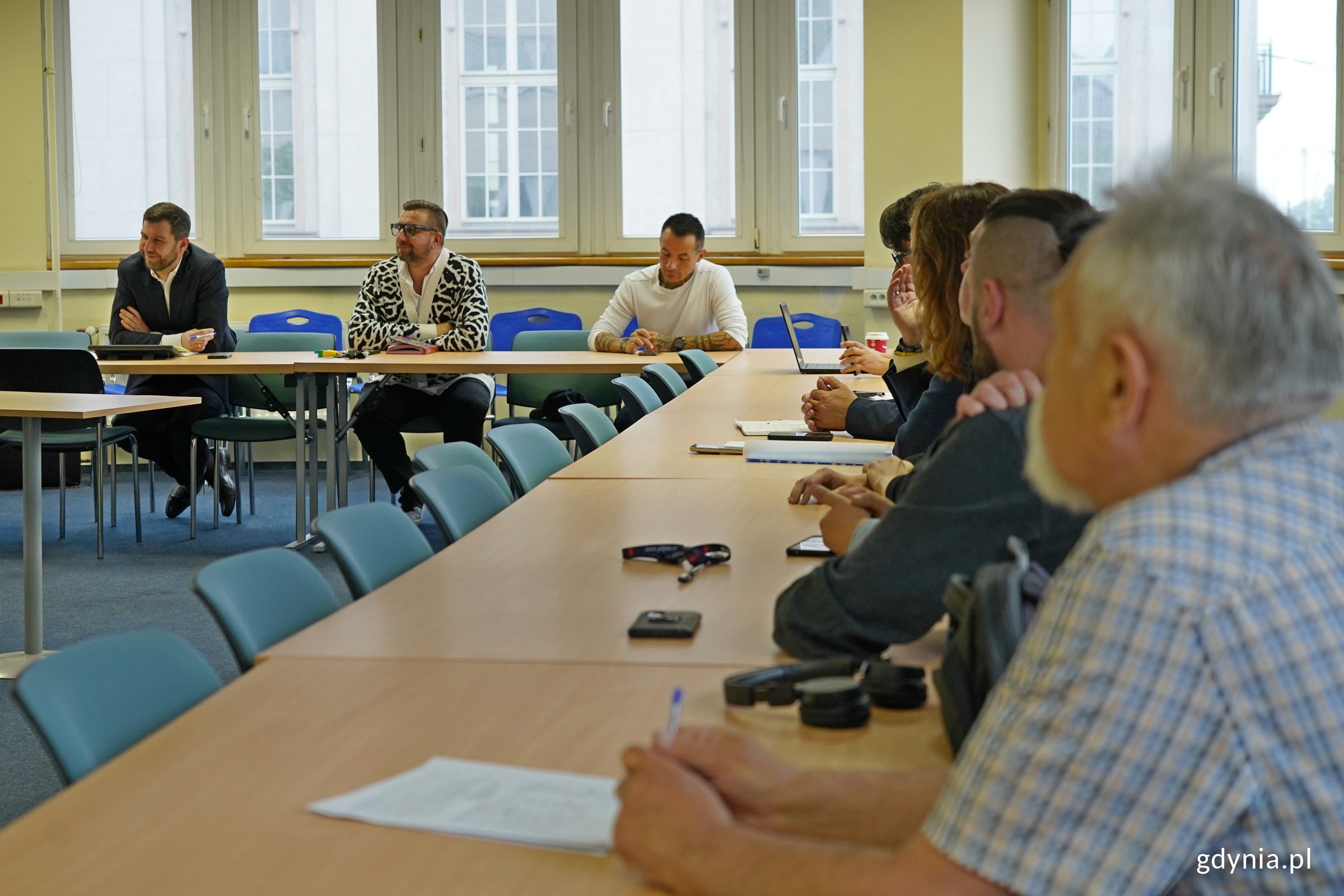 Sala w budynku Urzędu Miasta Gdyni, grupa os&oacute;b siedzi przy ustawionych w rzędzie stolikach