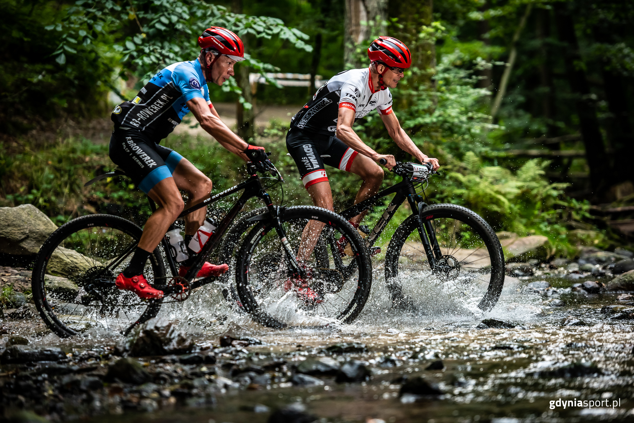 Dwaj zawodnicy na rowerach na leśnym odcinku trasy. Jadą po wodzie, kt&oacute;ra odpryskuje im spod opon. Mają na głowach czerwone kaski // fot. gdyniasport.pl