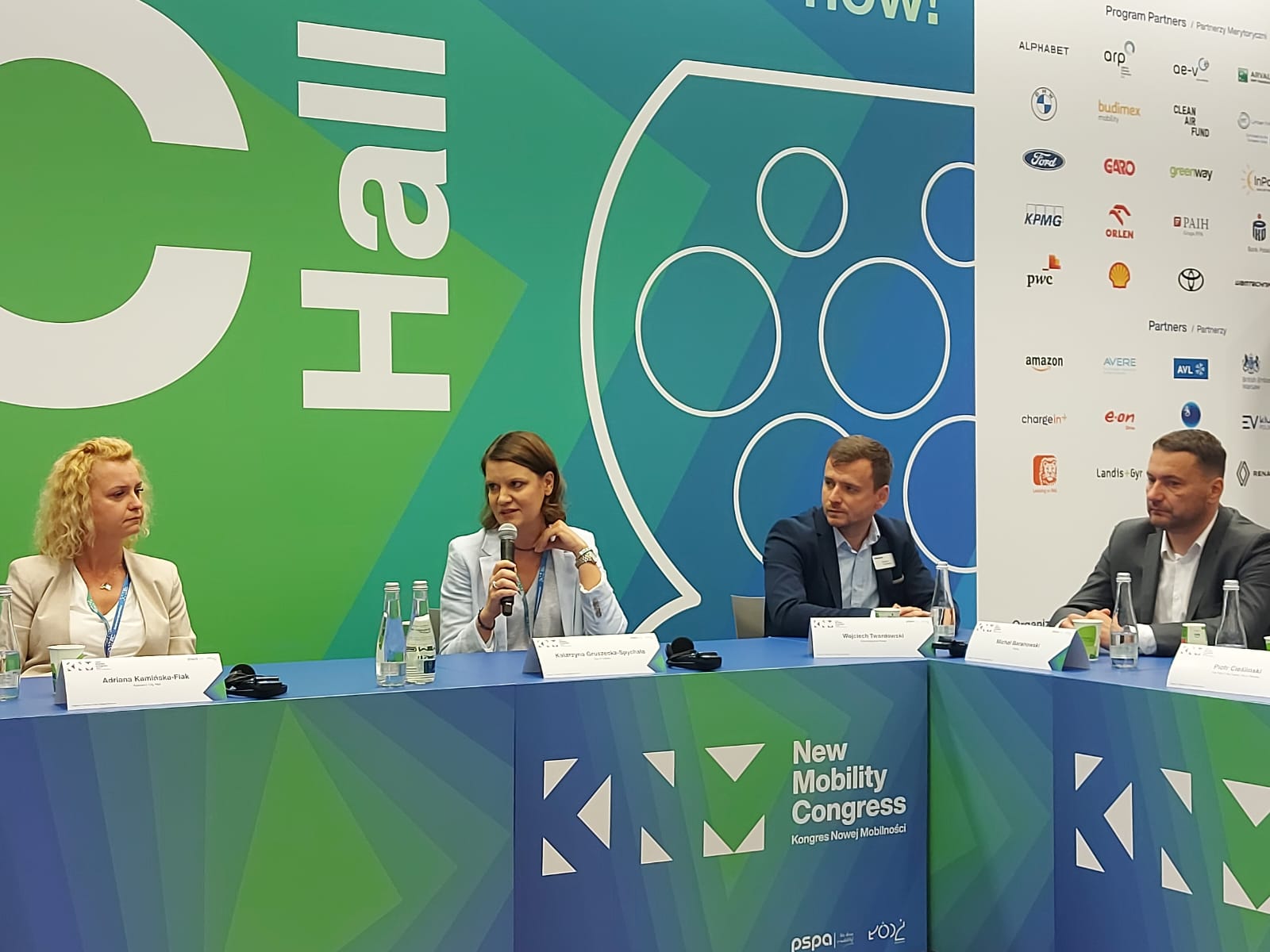 Sala konferencyjna, dekoracje tła, st&oacute;ł. Przy stole kilkoro prelegent&oacute;w, wypowiada się przez mikrofon Katarzyna Gruszecka-Spychała