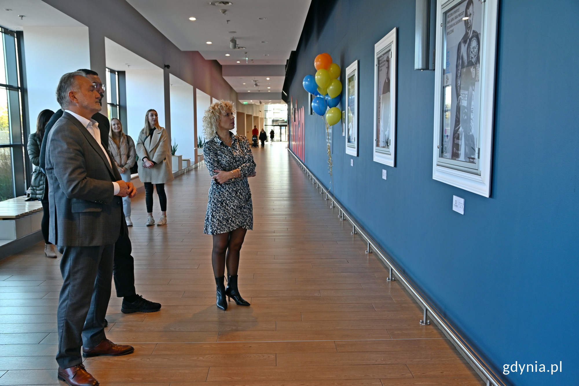 Od lewej: Wojciech Szczurek &ndash; prezydent Gdyni, Paweł Brach &ndash; dyrektor marketingu Centrum Riviera oraz Małgorzata Marmajewska &ndash; prezes fundacji &bdquo;Gdyński Most Nadziei&rdquo; i koordynatorka kampanii &bdquo;Odważni Wygrywają&rdquo; podczas wernisażu wystawy w Centrum Riviera. Za nimi inni uczestnicy wydarzenia. W tle hol i idący nim ludzie // fot. Magdalena Czernek