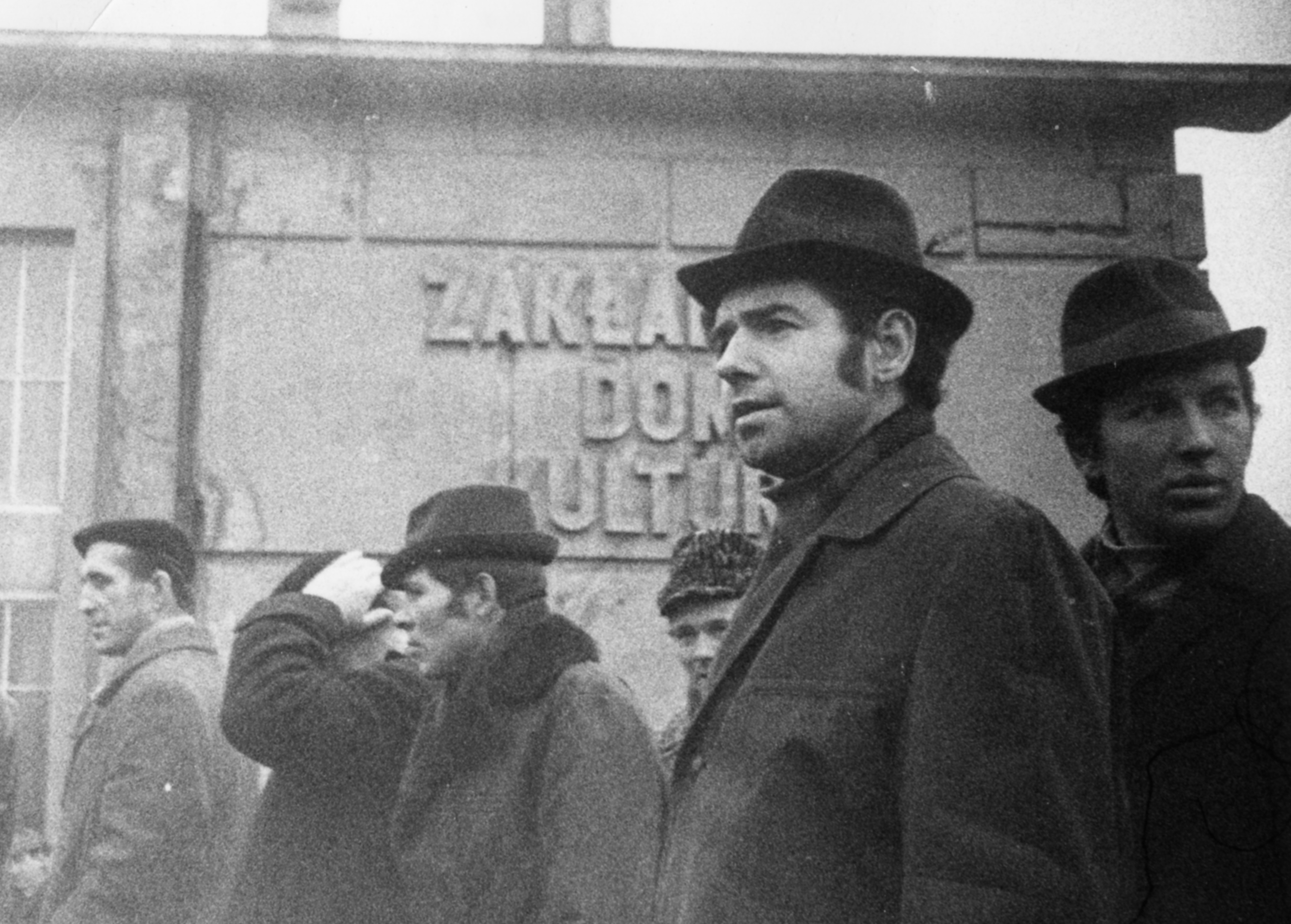 Mężczyźni przed budynkiem z widocznym na ścianie napisem "Zakładowy Dom Kultury" prawdopodobnie należącym do Stoczni im.Komuny Paryskiej w Gdyni. Budynek znajduje się przy ul.Polskiej // źr&oacute;dło: IPN