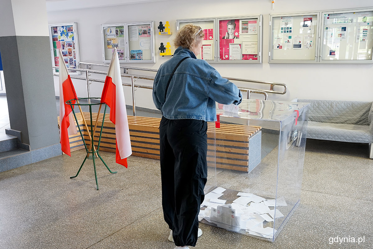 Hol szkoły w Gdyni, kobieta odwr&oacute;cona plecami wrzuca głos do urny wyborczej, w tle biało-czerwone flagi