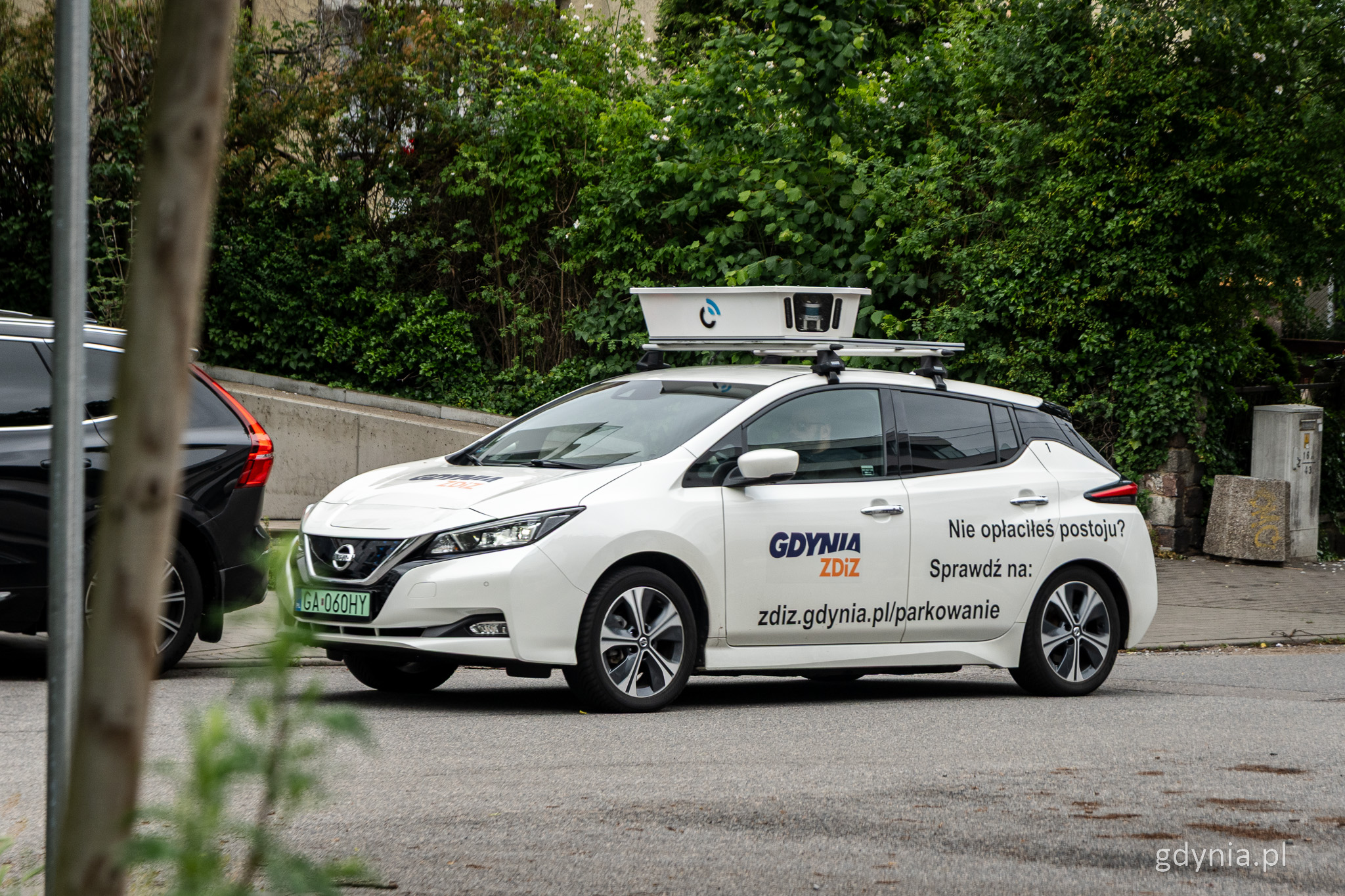 Ulica w Gdyni, biały samoch&oacute;d z logotypem ZDiZ w Gdyni, na dachu aparatura LIDAR do kontroli parkowania