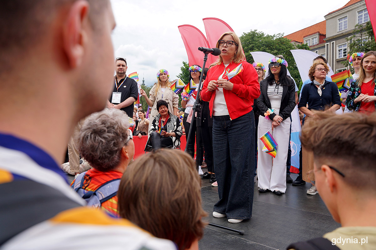 Scena plenerowa, tłum ludzi w kolorowych ubraniach, przy mikrofonie przemawia Oktawia Gorzeńska