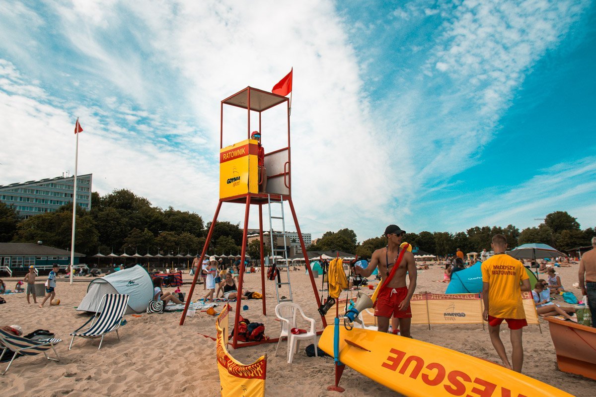 Plaża w Śr&oacute;dmieściu Gdyni, dzień, lato, ż&oacute;łta budka ratownik&oacute;w z drabiną, dookoła ludzie na plaży, w tle budynki