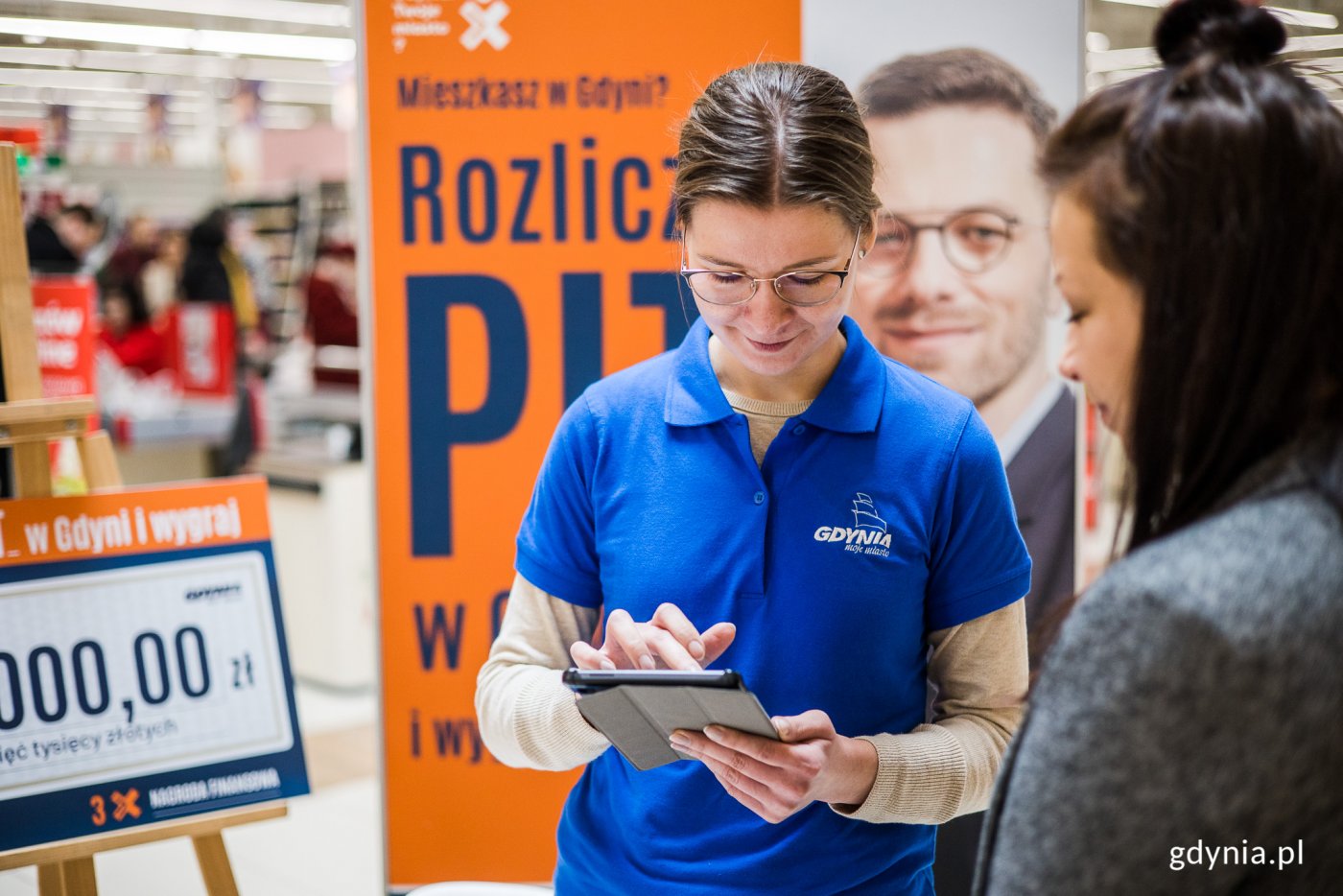 Kobieta w niebieskiej koszulce trzyma tablet, obok druga kobieta. W tle ścianki promujące kampanię Rozlicz PIT w Gdyni w pomarańczowym kolorze, w tle kasy hipermarketu w centrum handlowym Riviera w Gdyni