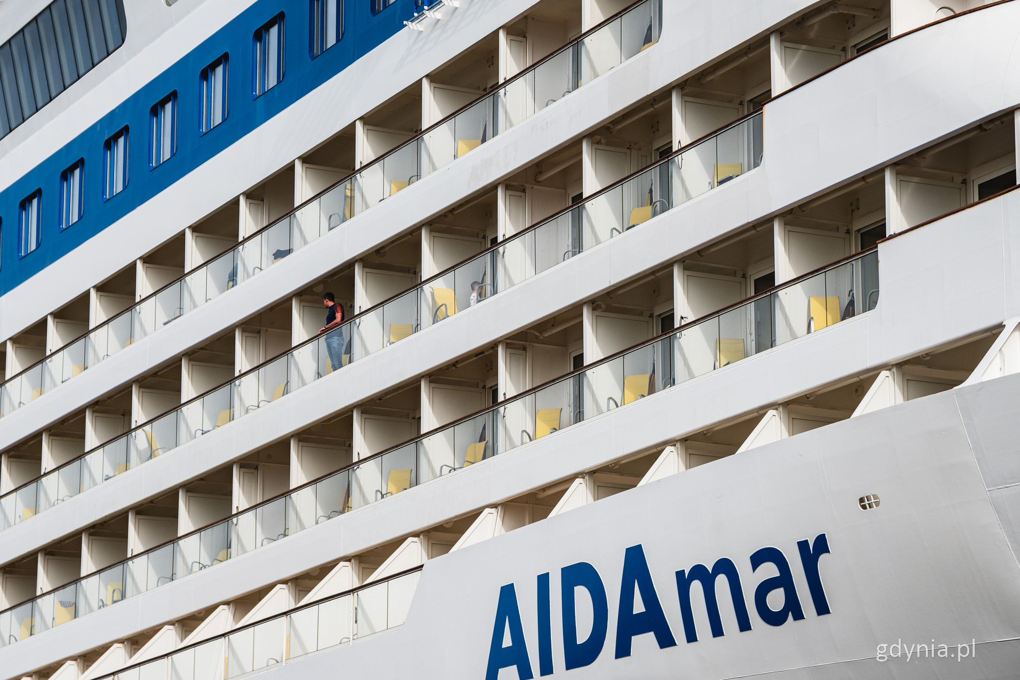 Burta statku wycieczkowego w białym kolorze, balkony, krzesła, kilka os&oacute;b ogląda nabrzeże na balkonach