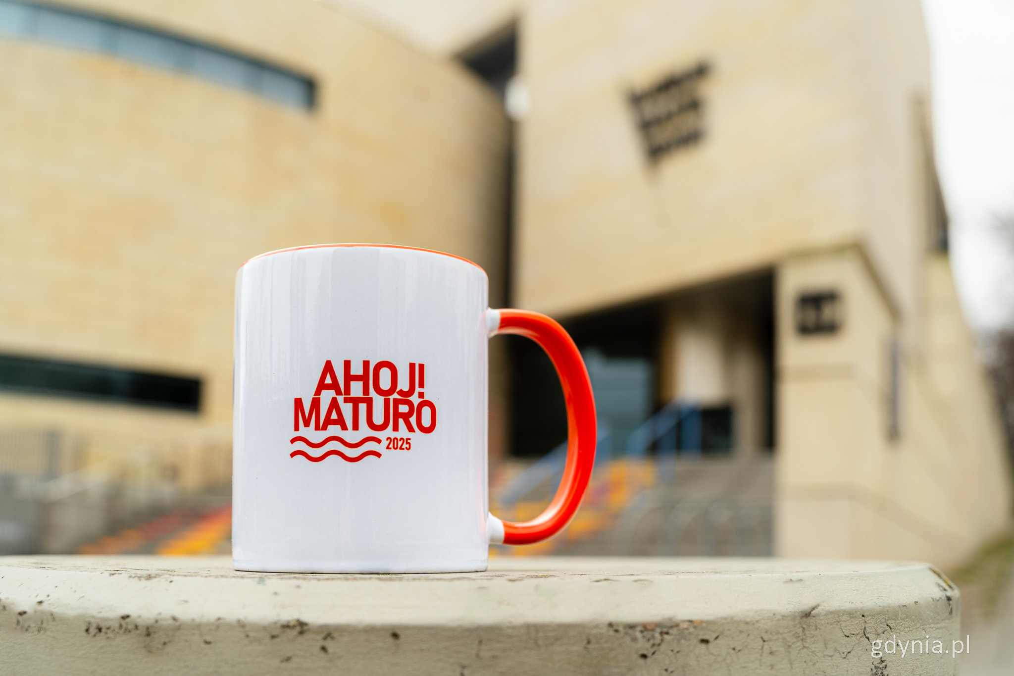 Biały kubek z pomarańczowym napisem "Ahoj! Matura", w tle muzeum miasta Gdyni i schody
