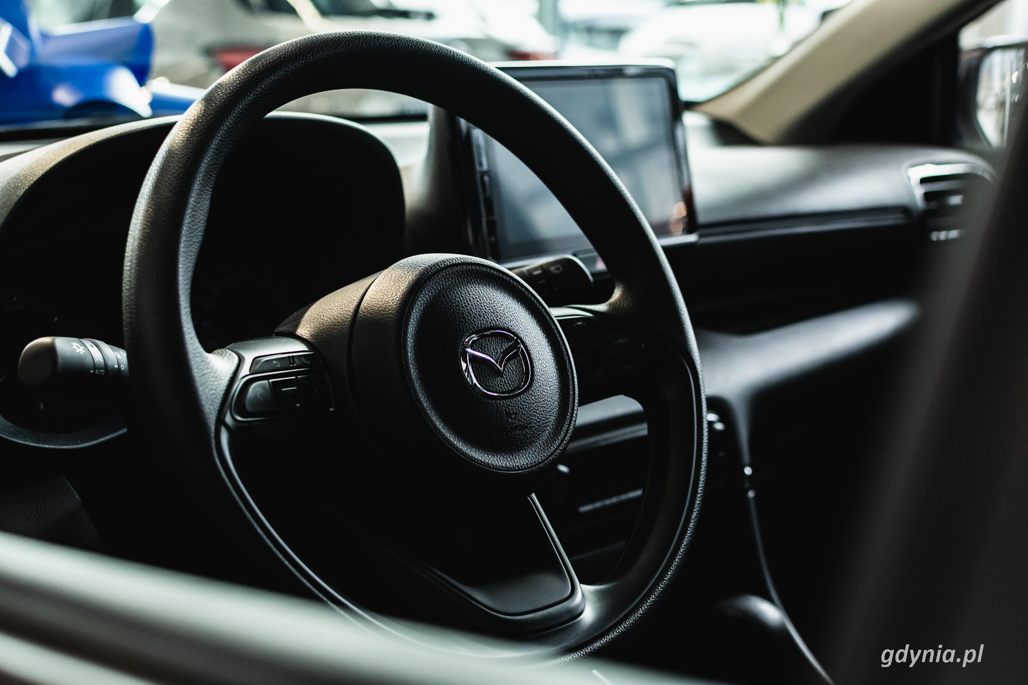 Wnętrze samochodu, widoczna kierownica z logo Mazdy i ciemna deska rozdzielcza, w tle wnętrze salonu samochodowego