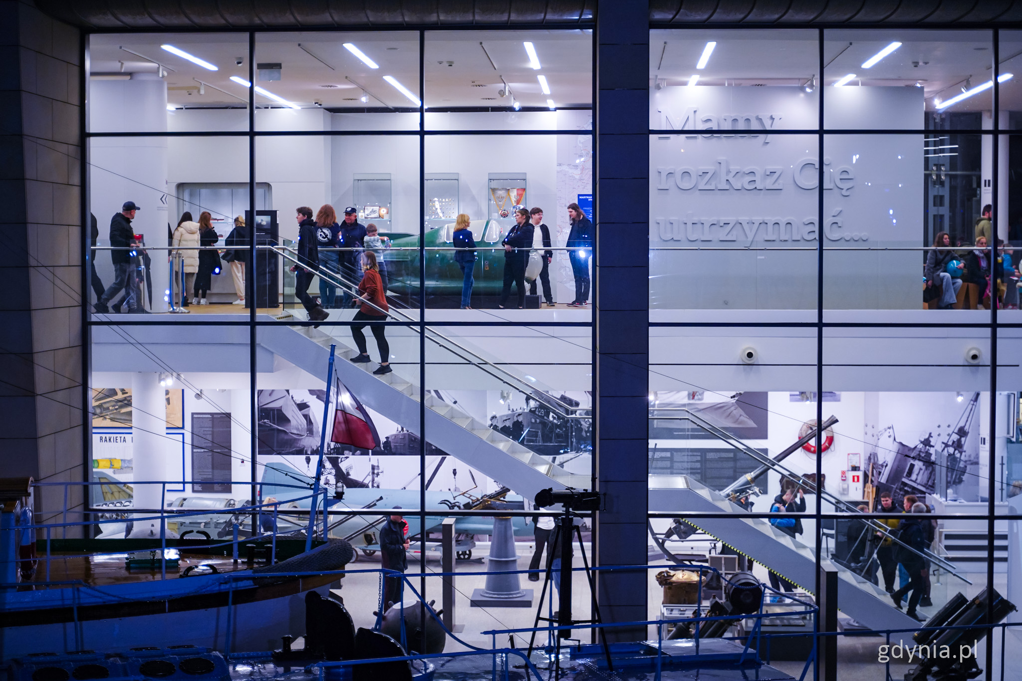 Przeszklona elewacja Muzeum Marynarki Wojennej, noc, widać wystawę i ludzi zwiedzających wystawę