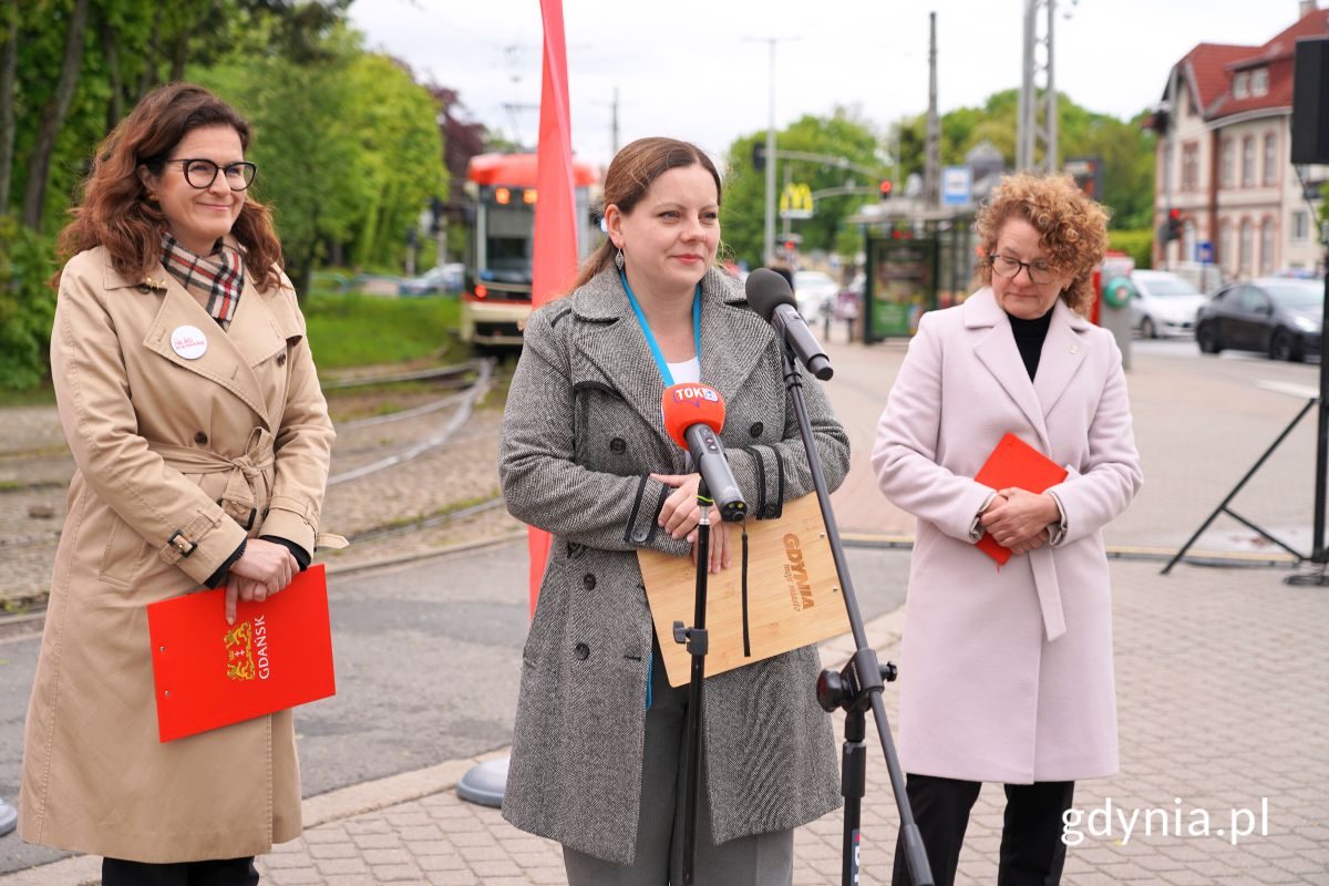 Prezydent Gdańska Aleksandra Dulkiewicz, prezydent Gdyni Aleksandra Kosiorek oraz prezydentka Sopotu Magdalena Czarzyńska-Jachim stojące przy mikrofonach na tle tramwaju oraz polskich flag
