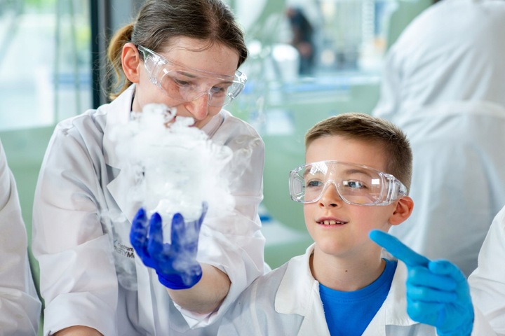 Edukatorka i chłopiec w białych fartuchach w Centrum Nauki Experyment Gdyni. Oboje mają nałożone przezroczyste gogle ochronne. Edukatorka trzyma w ręku naczynie, z kt&oacute;rego wydobywa się biały dym // fot. materiały prasowe