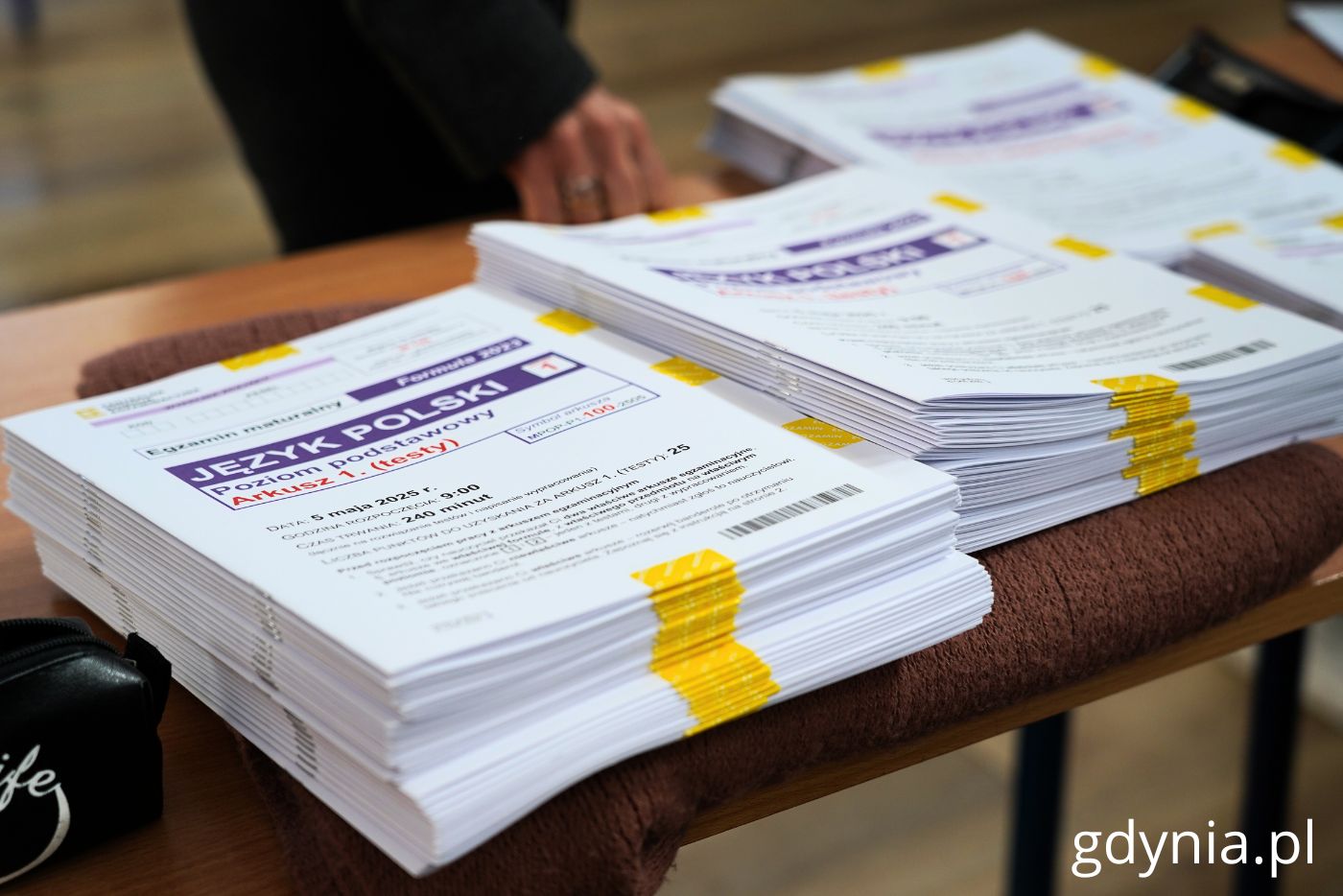 St&oacute;ł w szkole, na blacie stos egzamin&oacute;w maturalnych, na froncie duży napis Język Polski