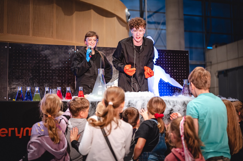 Noc Naukowc&oacute;w w Centrum Nauki Experyment, edycja 2022. Dzieci stoją przed sceną, na kt&oacute;rej stoi dw&oacute;jka naukowc&oacute;w, kobieta i mężczyzna. 