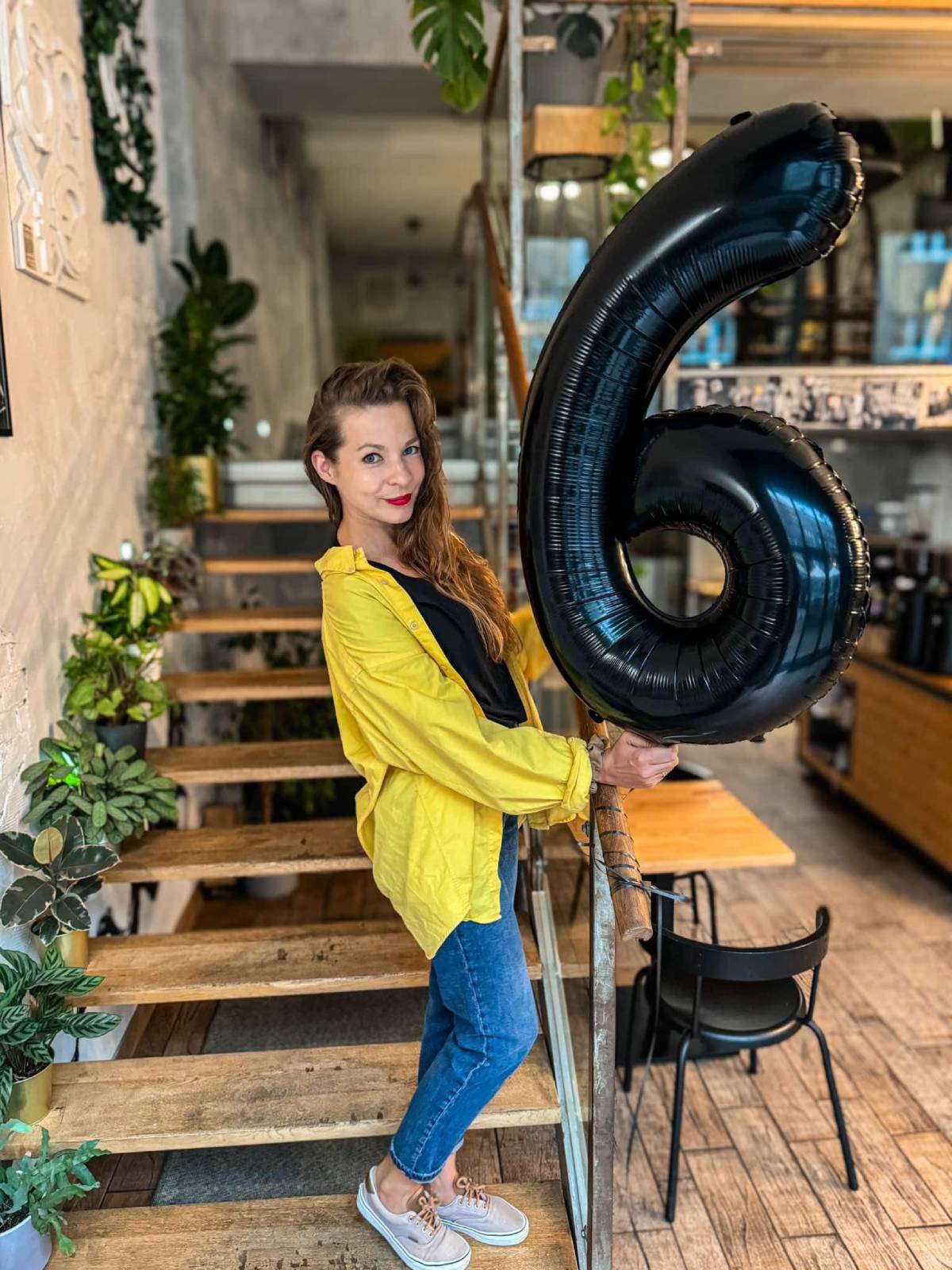 Kobieta w ż&oacute;łtej koszuli i jeansach stojąca na drewnianych schodach wewnątrz kawiarni i trzymająca czarny balon w kształcie cyfry 6