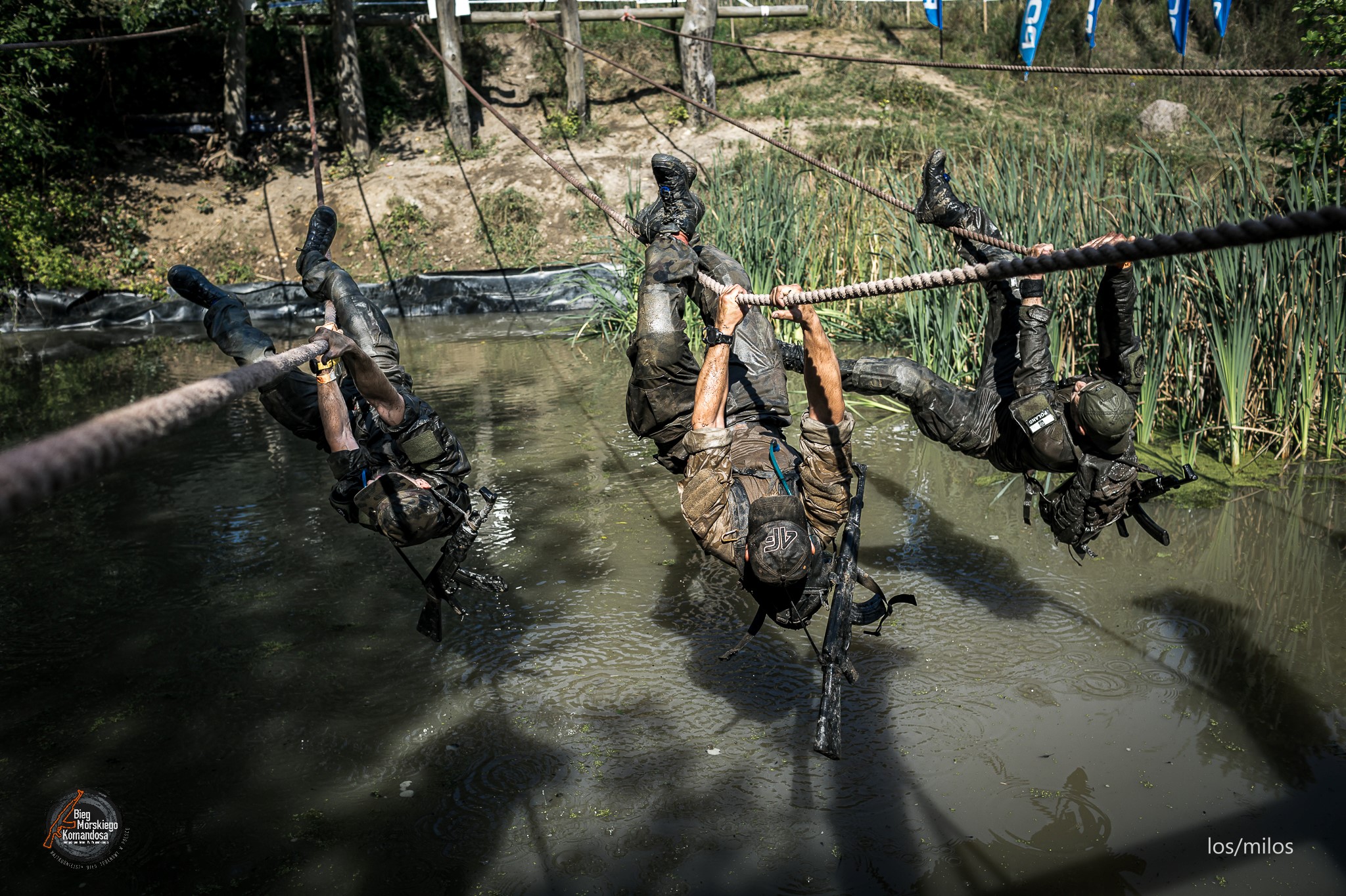 Na obrazku znajduje sie troje uczestnik&oacute;w Biegu Morskiego Komandosa, kt&oacute;rzy trzymają się liny nad tafla wody. W tle trawa, krzaki i drzewa.