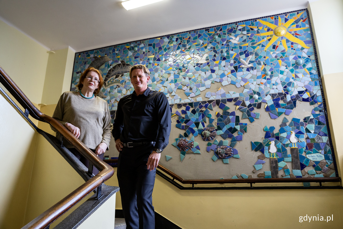 Kobieta i mężczyzna stoją na p&oacute;łpiętrze klatki schodowej, za nimi mozaika na ścianie przedstawiająca morze. 