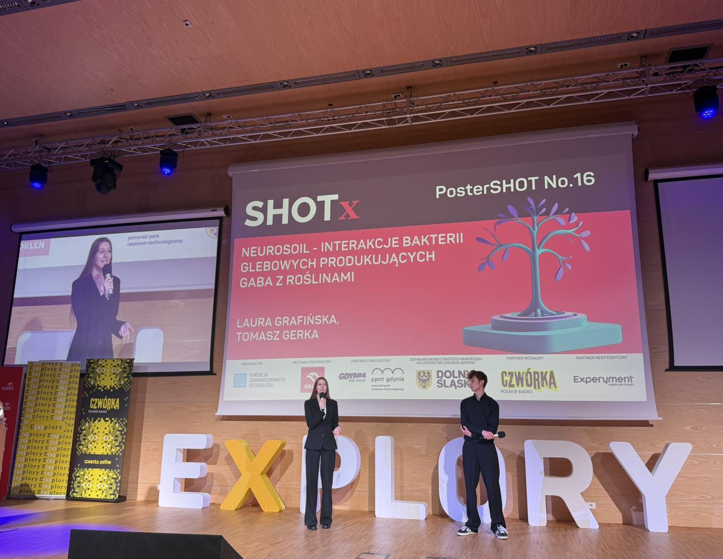 Scena na sali konferencyjnej w PPNT Gdynia, prezentacja projektu na ekranie, dziewczyna i chłopak prezentują na scenie, m&oacute;wią do mikrofon&oacute;w