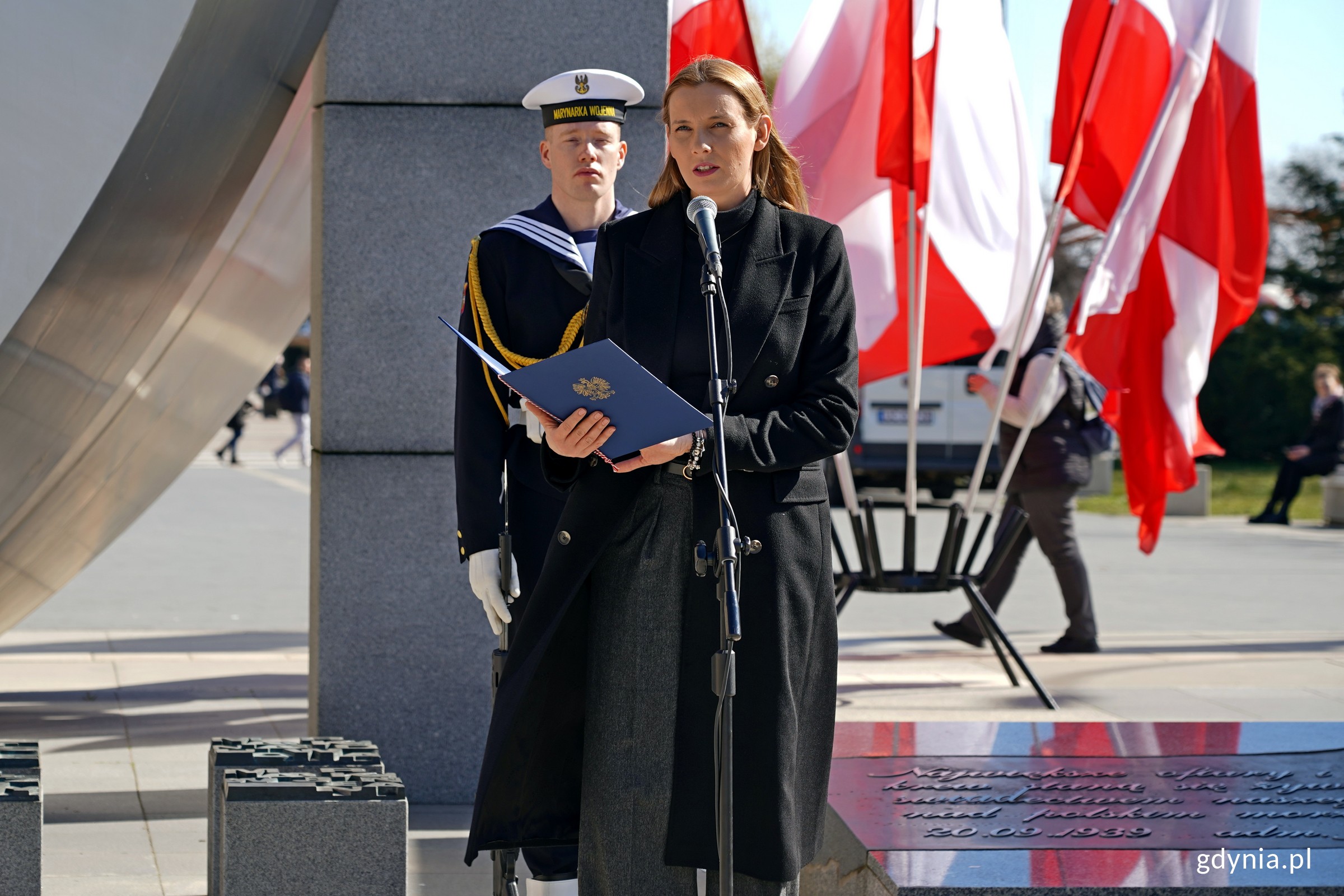 Anna Chojnowska, naczelniczka Biura Prezydenta, odczytująca list od prezydent Aleksandry Kosiorek podczas uroczystości z okazji 80. rocznicy zakończenia walk o Gdynię. W tle marynarz stojący pod pomnikiem Polski Morskiej i powiewające na wietrze biało-czerwone flagi