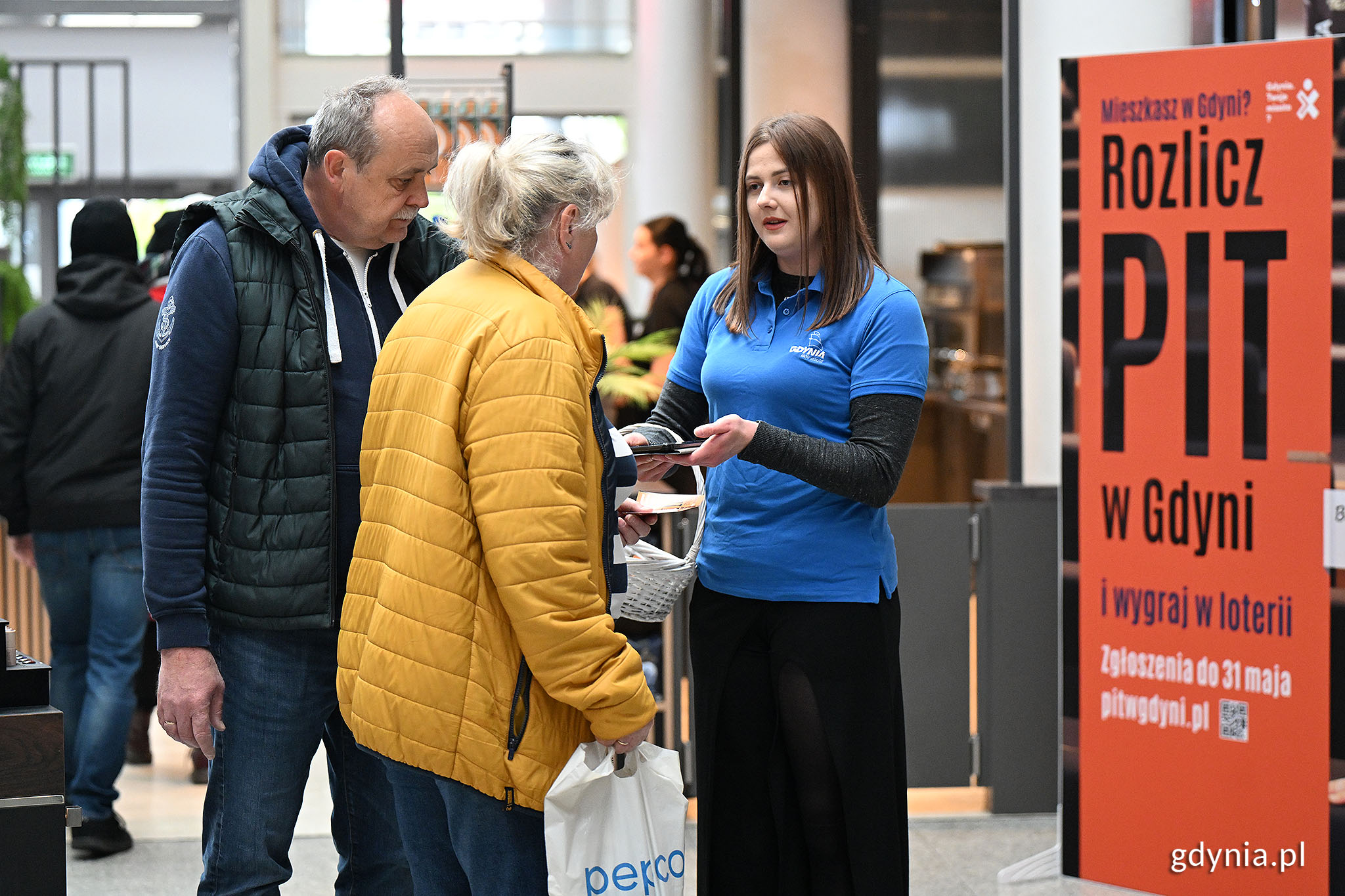 CH Batory, para rozmawia z kobietą w niebieskiej koszulce, kt&oacute;ra trzyma tablet, w tle ekspozycja z logo "Rozlicz PIT w Gdyni"