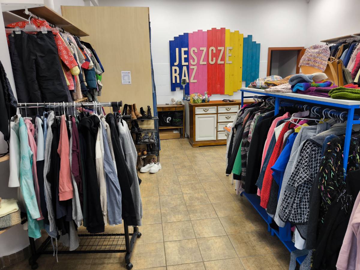 Wieszaki z kolorowymi ubraniami ustawione wewnątrz sklepu, w tle widoczna biała ściana i nazwa sklepu