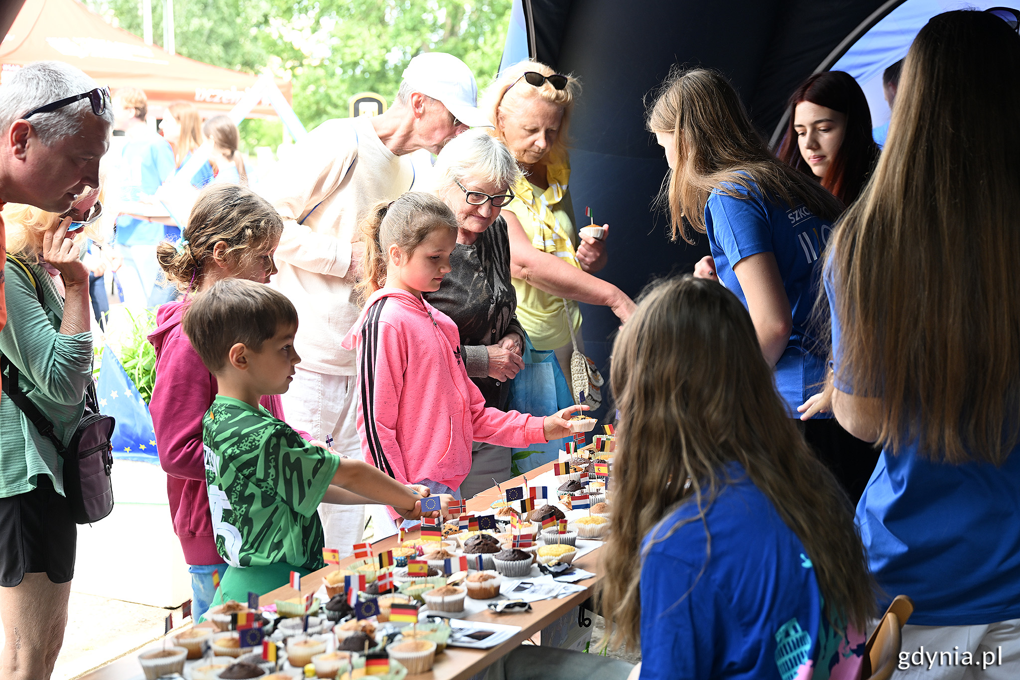 Namiot na Placu Wolnej Ukrainy, kilka kobiet w niebieskich koszulkach oraz odwiedzający stoją przy stoliku, na stoliku ciastka