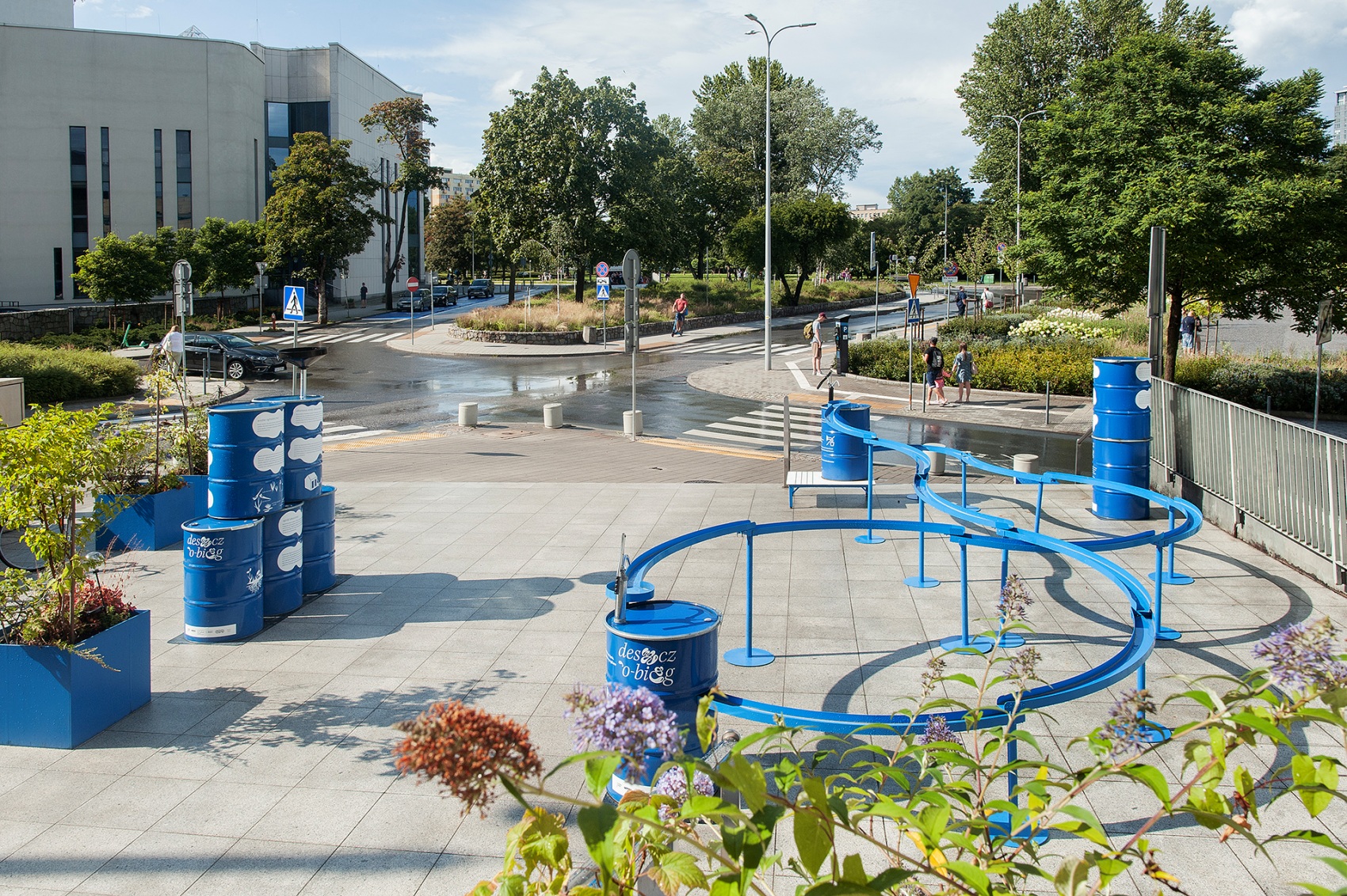 Plac przed Muzeum Miasta Gdyni, lato, słoneczny dzień, niebieska instalacja artystyczna, beczki rury, kwiaty, zieleń