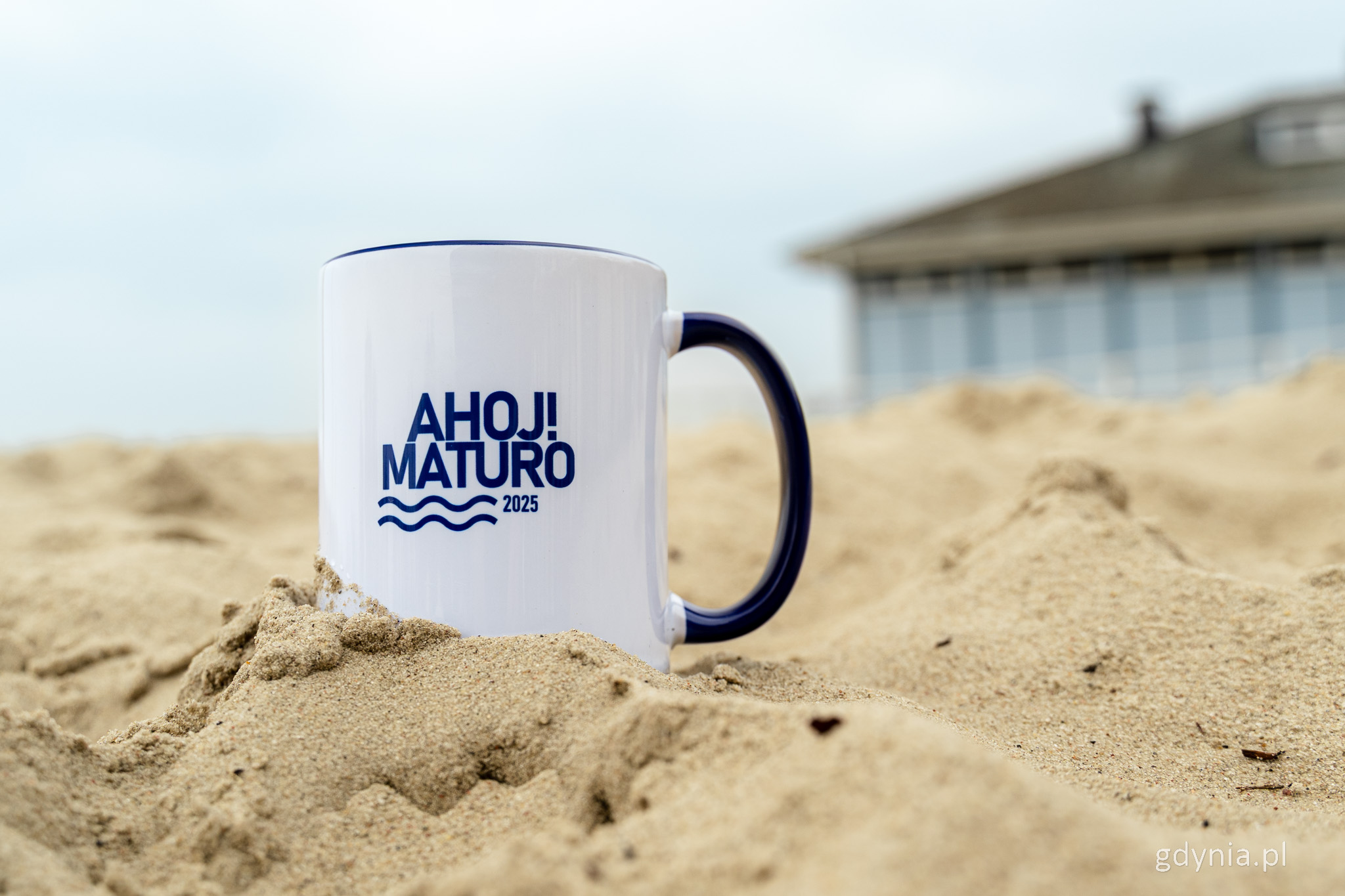 Biały kubek z ciemnoniebieskim napisem "Ahoj! Maturo" na tle plaży i lokalu Contrast w Gdyni