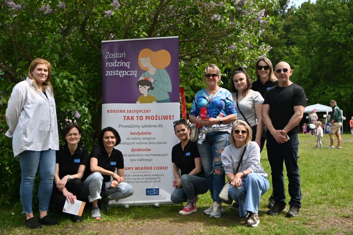 Zdjęcie przedstawia grupę ludzi w r&oacute;żnym wieku, przy bannerze promujący rodzicielstwo zastępcze w Gdyni
