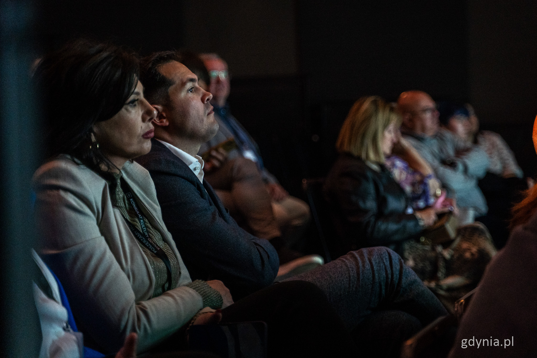 Widownia w sali Konsulatu Kultury, przygaszone światło, osoby siedzą na krzesłach, mężczyzna i kobieta obserwują scenę