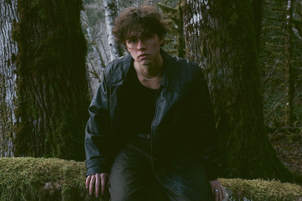 na zdjęciu młody mężczyzna z kręconymi włosami stoi w lesie