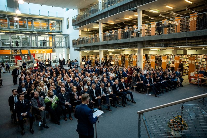 Sala konferencyjna, tłum osób siedzących na krzesłach, biblioteczne regały w tle, na środku stoi przemawiający przez mikrofon mężczyzna