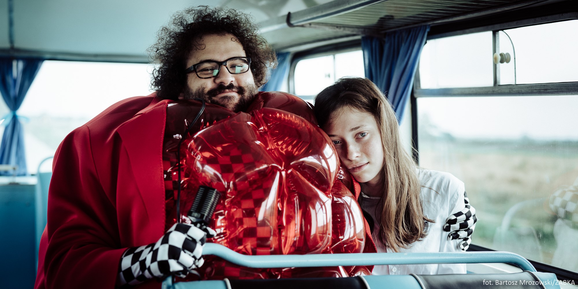 Kadr z filmu "Alicja i Żabka". Mężczyzna i dziewczyna siedzą obok siebie w autobusie. Mężczyzna ma na sobie czerwony dmuchany kostium i okulary. Dziewczyna przytula się do niego // fot. Bartosz Mrozowski