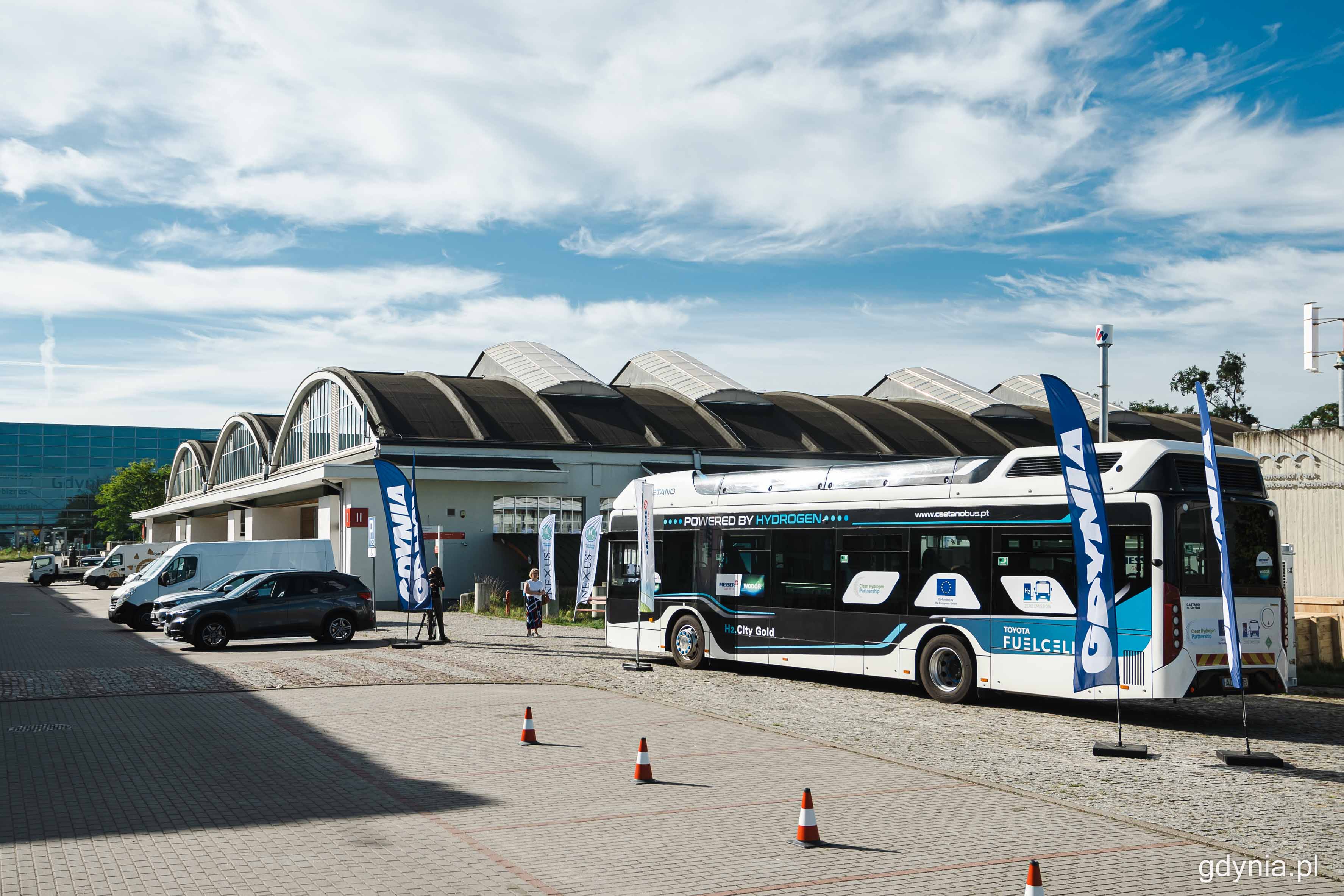 Plac przed PPNT Gdynia, słoneczny dzień, samochody na parkingu, w tle budynek Experymentu z napisem "Gdynia", drogą jedzie autobus wodorowy z napisem "Gdynia w klimacie" na wyświetlaczu